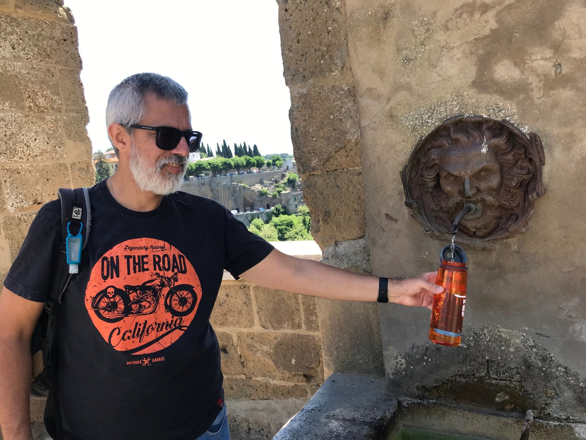 Uma pessoa de óculos escuros pegando água em uma antiga fonte na Itália