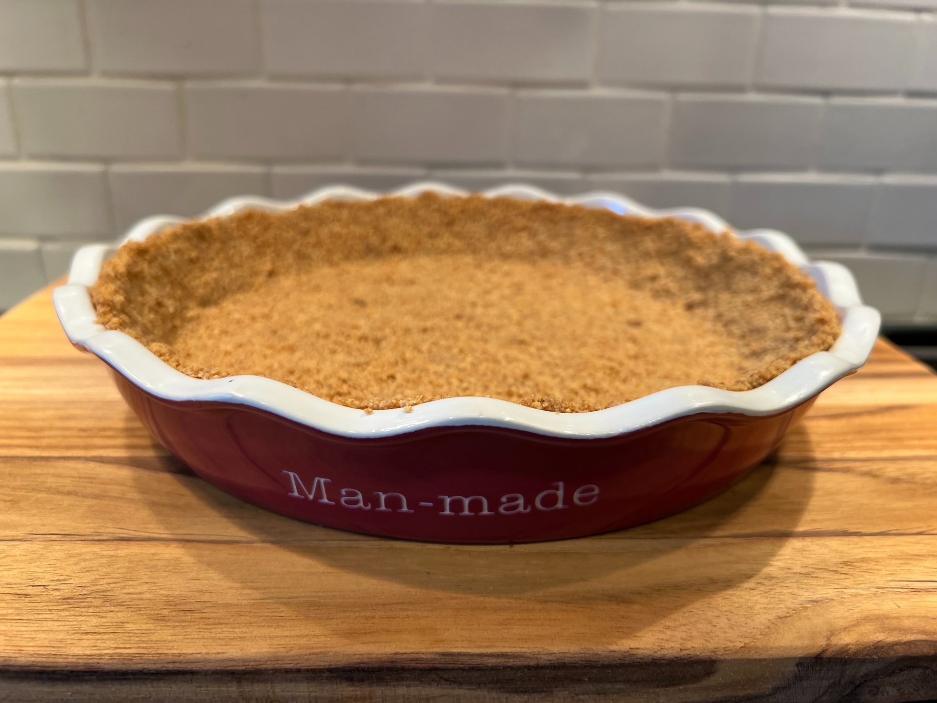 A homemade graham cracker pie crust pressed into a red ceramic pie dish on a wooden cutting board.