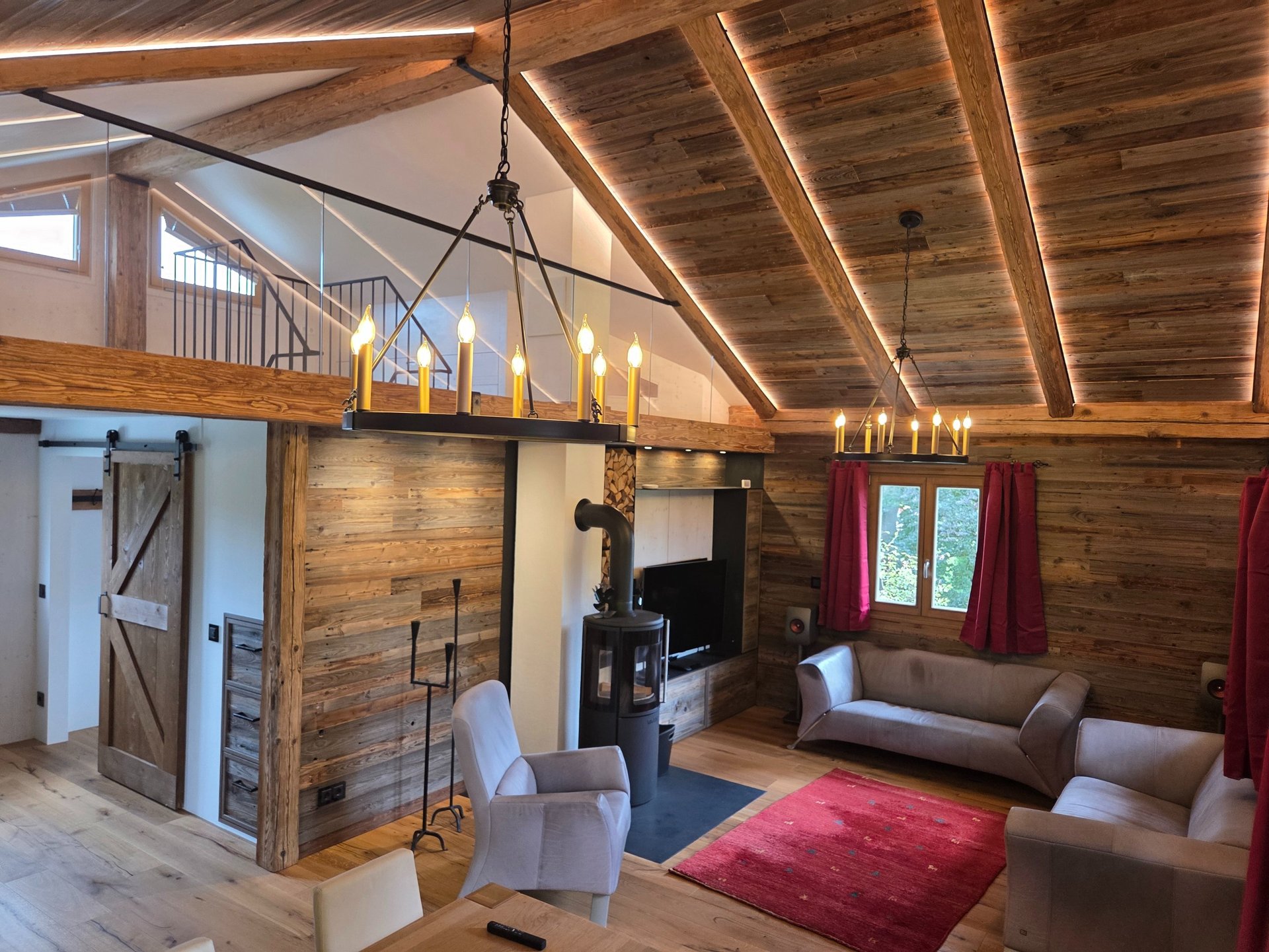 Großzügiges Wohnzimmer mit offenem Altholz-Dachstuhl, Galerie mit Glasgeländer, zwei Kronleuchtern und Kaminofen.