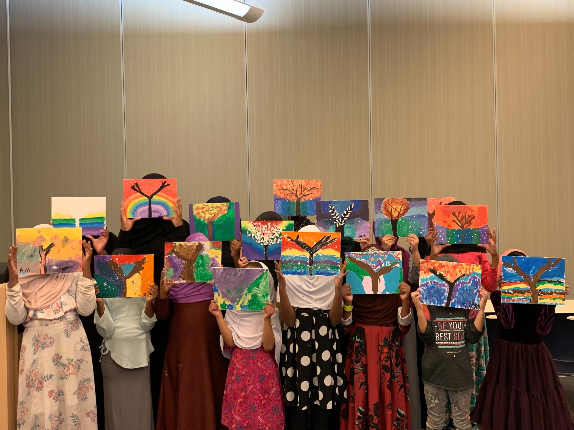 a group of Muslim girls standing in rows, holding colorful tree paintings up, hiding their faces