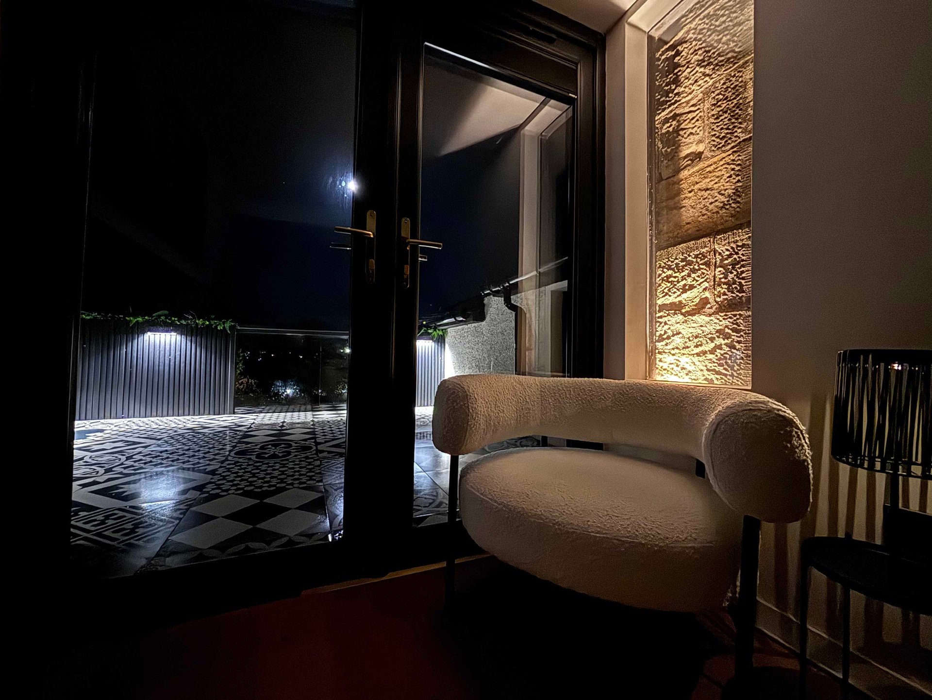 Modern white boucle armchair next to a black glass door overlooking a moonlit patio with geometric tiles.