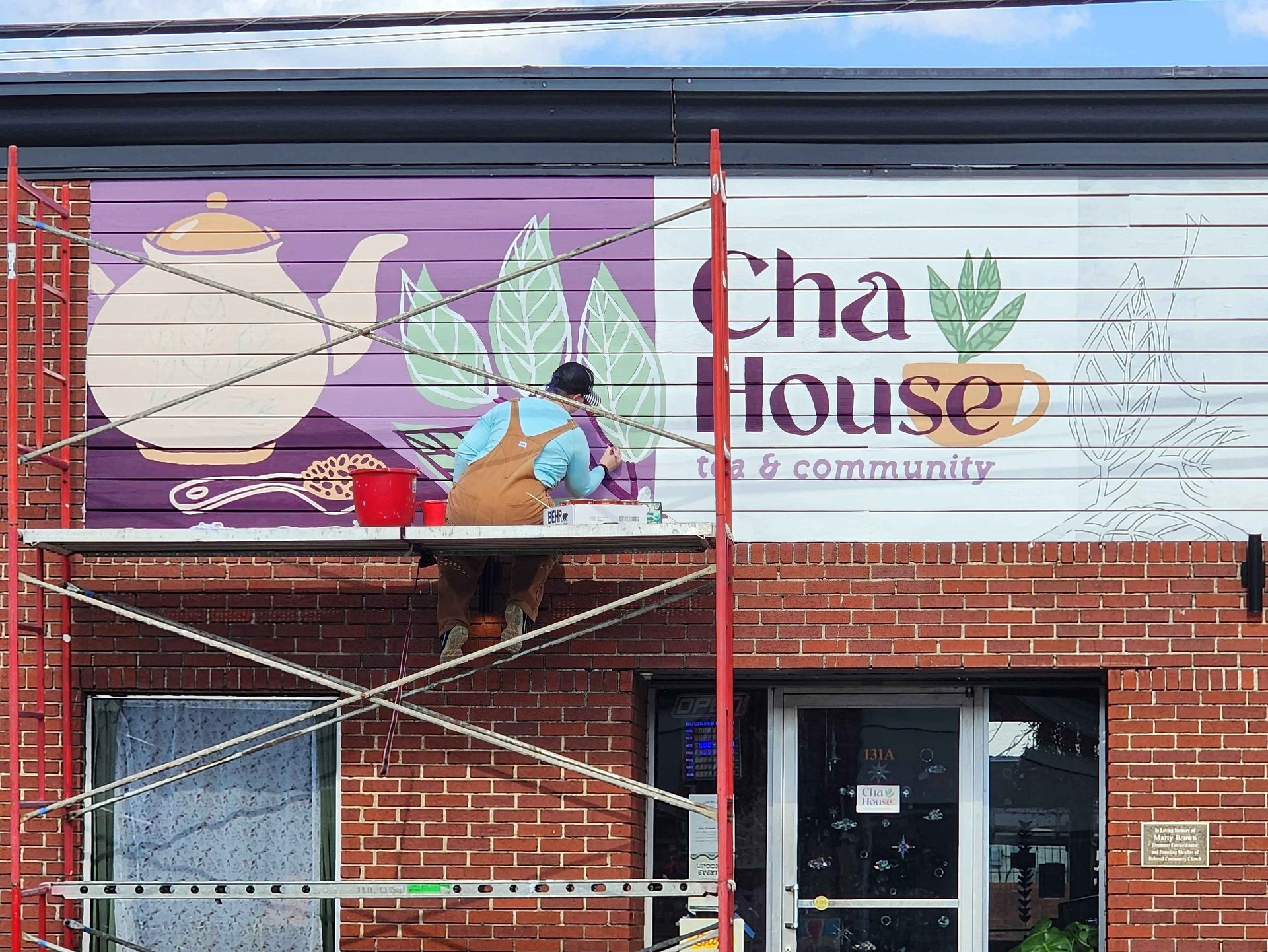 Artist on scaffolding painting a tea house mural with teapot and leaf graphics.