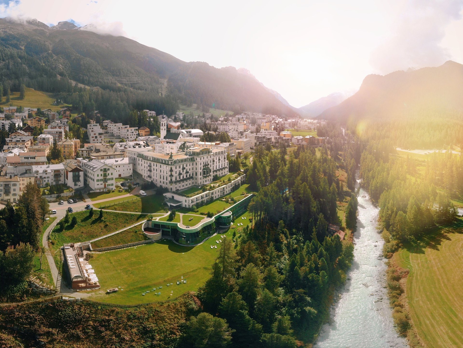 Aerial view of a luxury hotel and alpine village in a sunlit Swiss mountain valley next to a river.