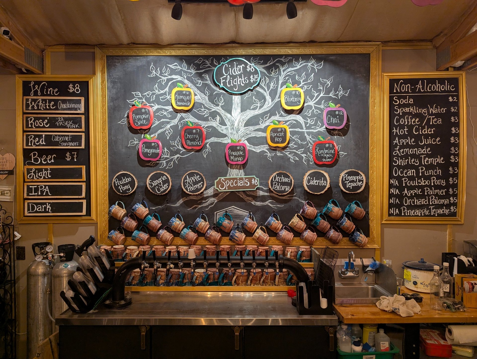 A chalkboard bar menu featuring cider flights, wine, beer, and non-alcoholic drinks above a row of tap handles.