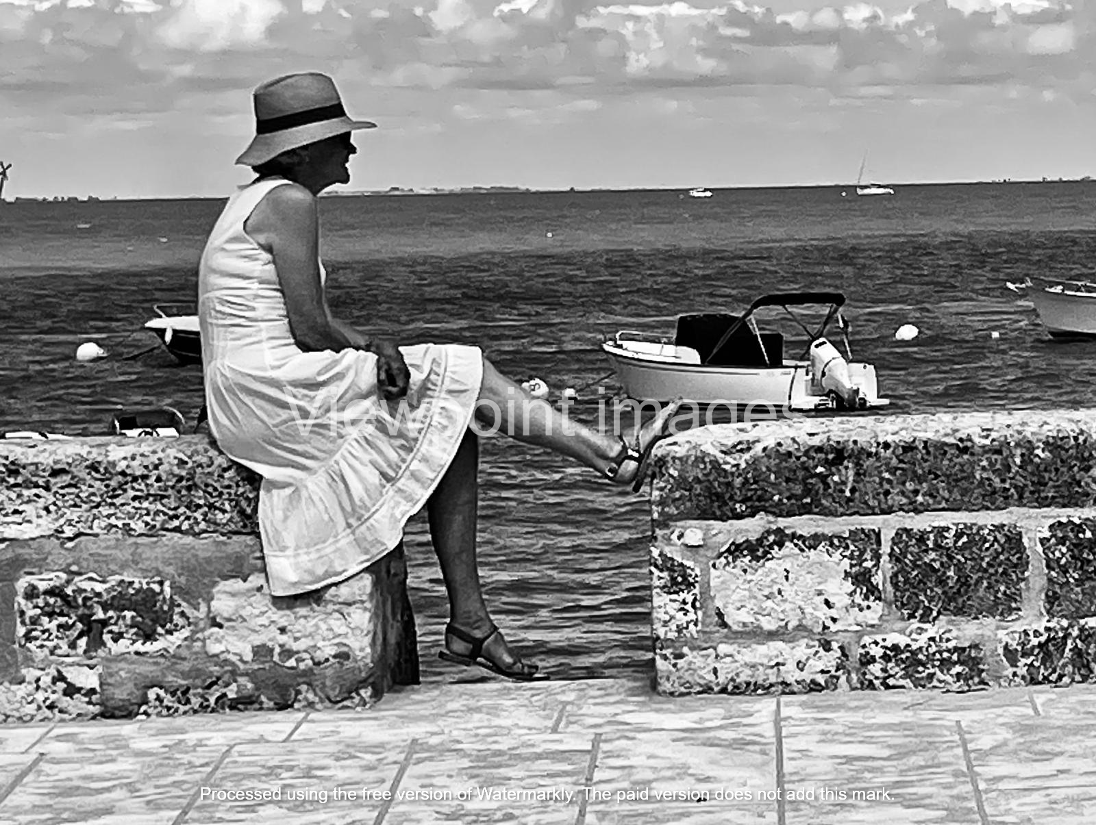 Black and white photo of a woman in a sun hat and white dress sitting on a stone wall by the sea.