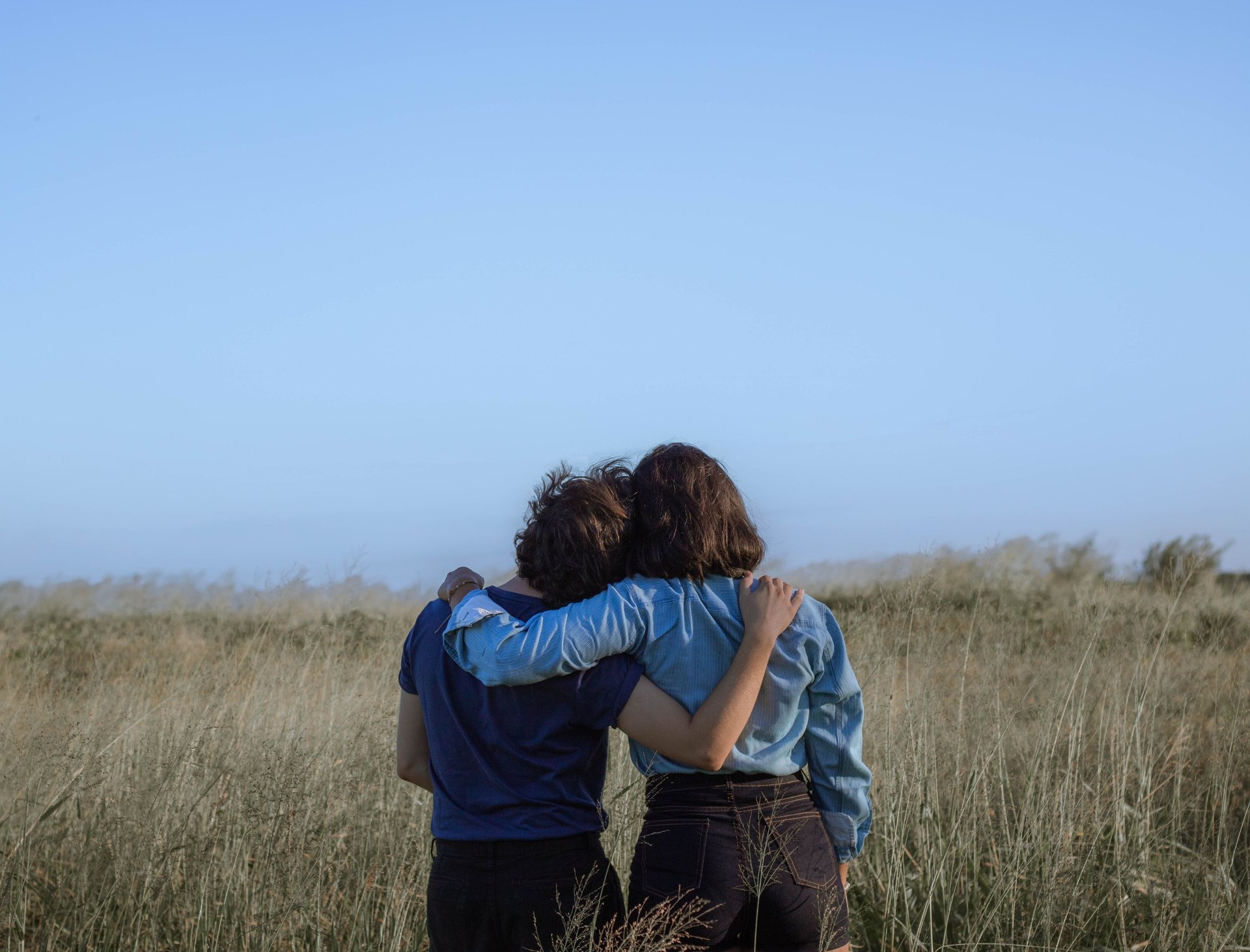 Two friends hugging and looking at the horizon in a grassy field under a clear blue sky.