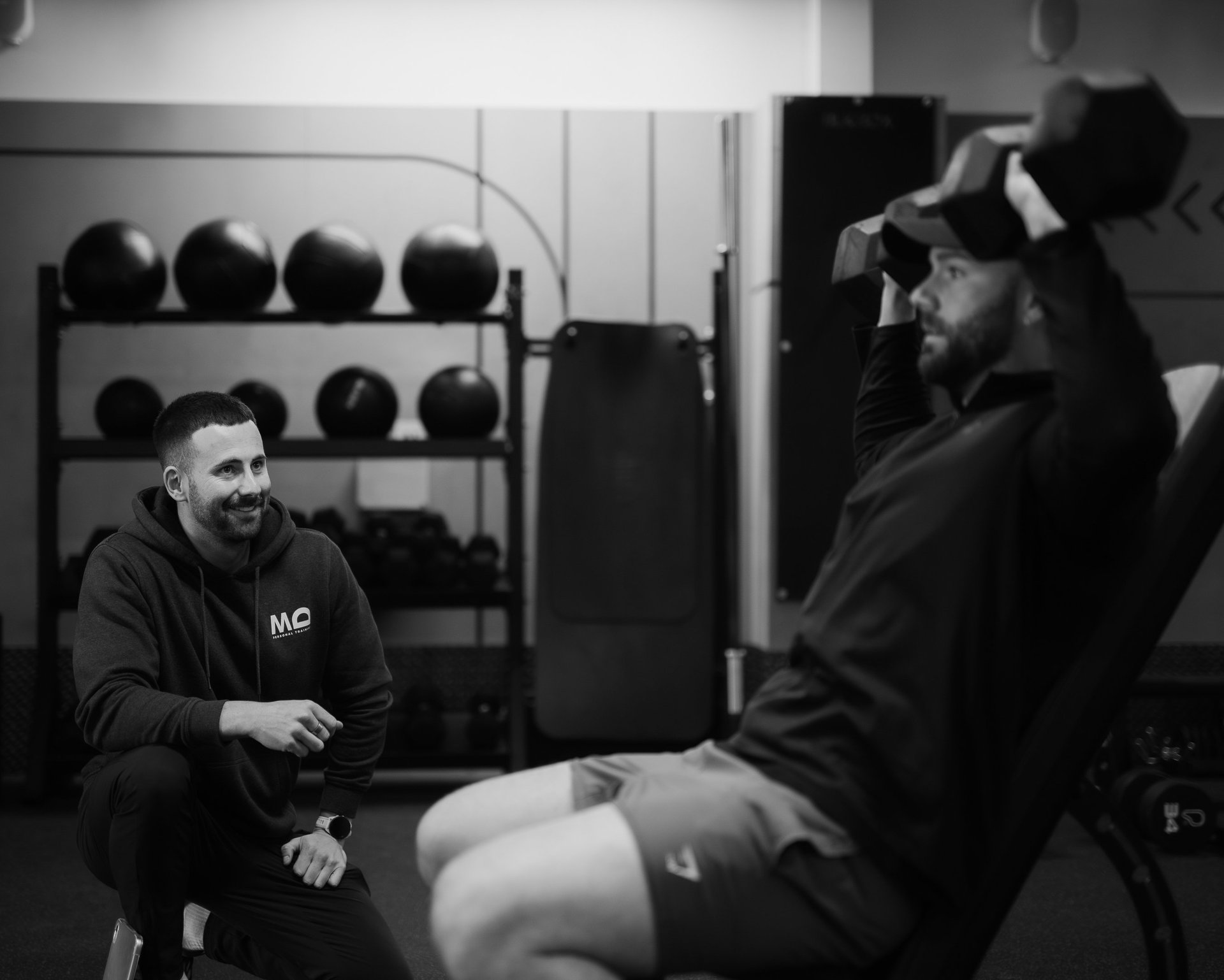 Trainer observing client’s seated dumbbell shoulder press in Leamington Spa gym, highlighting coaching and strength training.