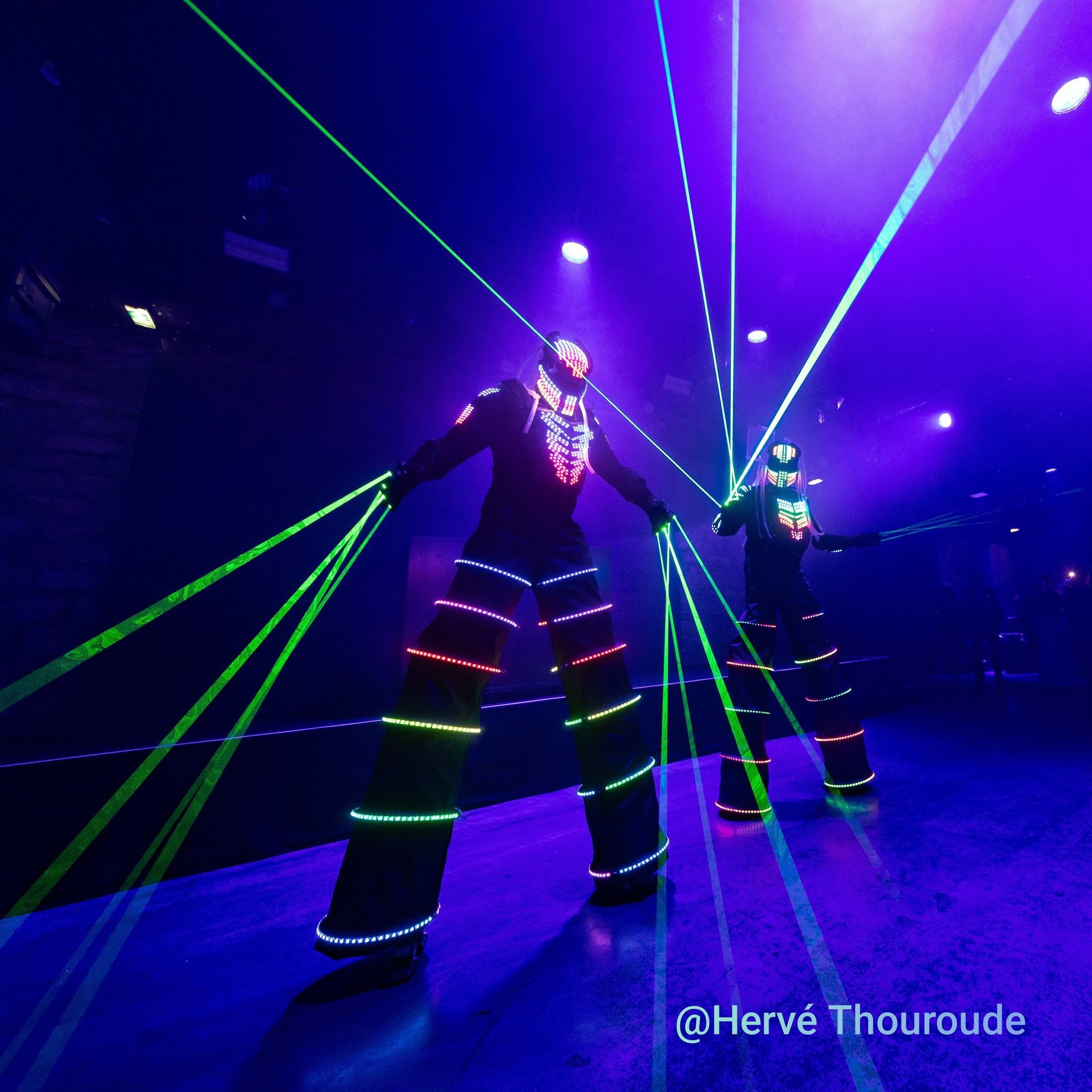 Performers on stilts in LED light suits and laser gloves at a high-tech neon nightclub event.
