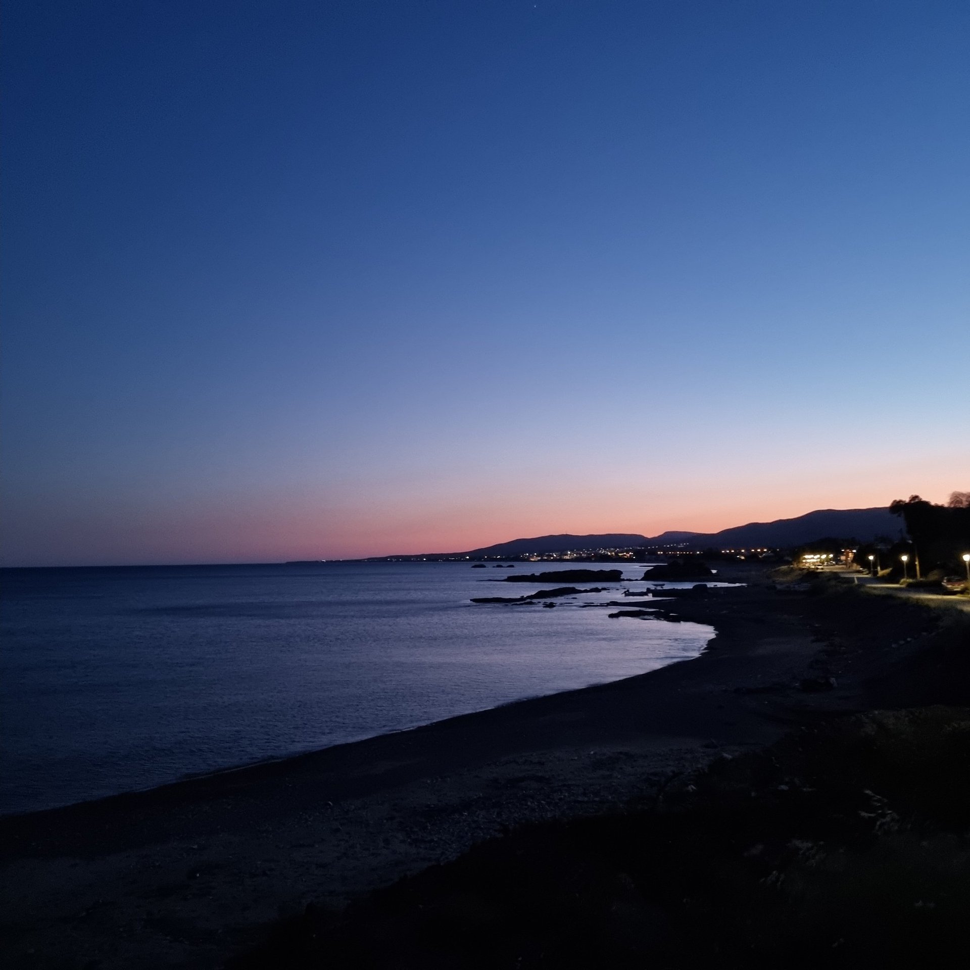 A scenic dusk view of a dark sandy beach coastline with distant city lights and a purple sunset horizon.