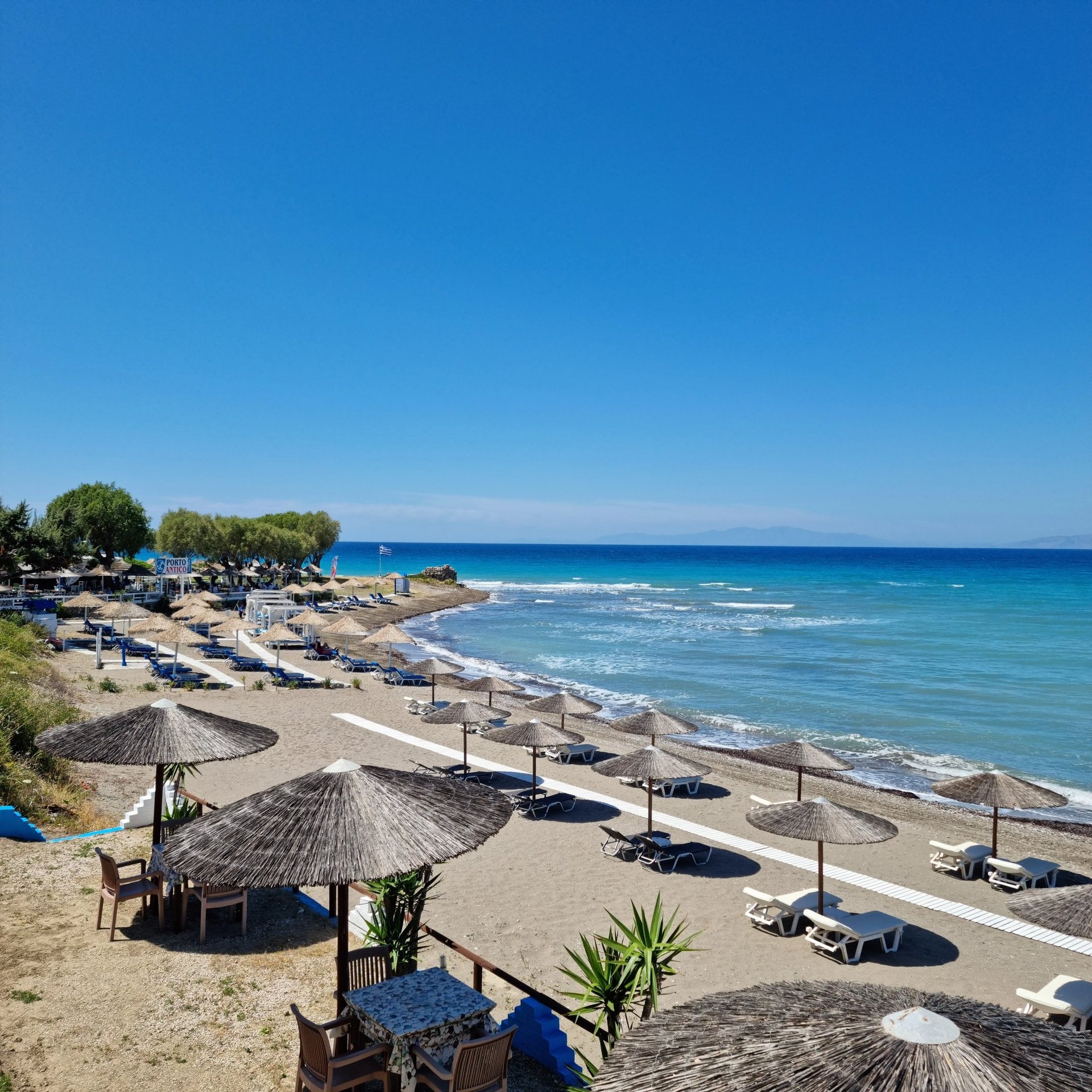 Sunny beach resort in Rhodes with straw umbrellas and lounge chairs by the blue Aegean Sea.