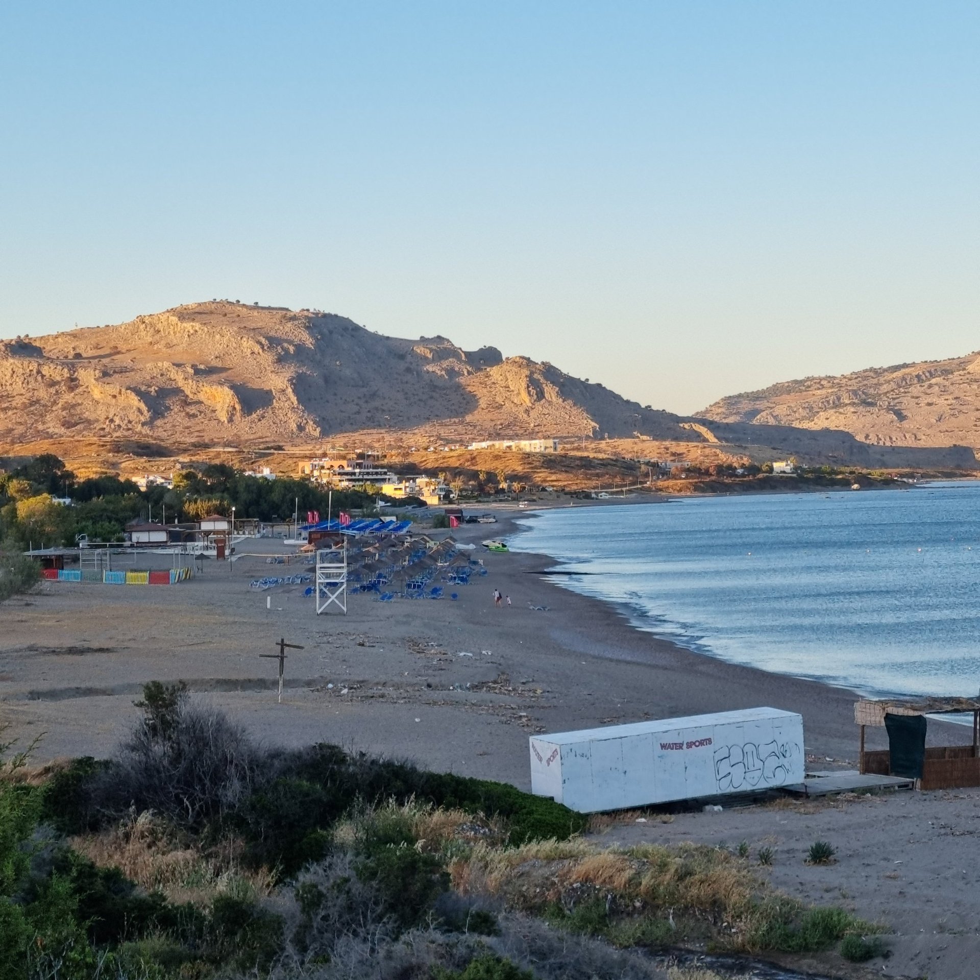 Panoramic view of a sandy Mediterranean beach with sunbeds, blue sea, and rocky mountains at sunset.