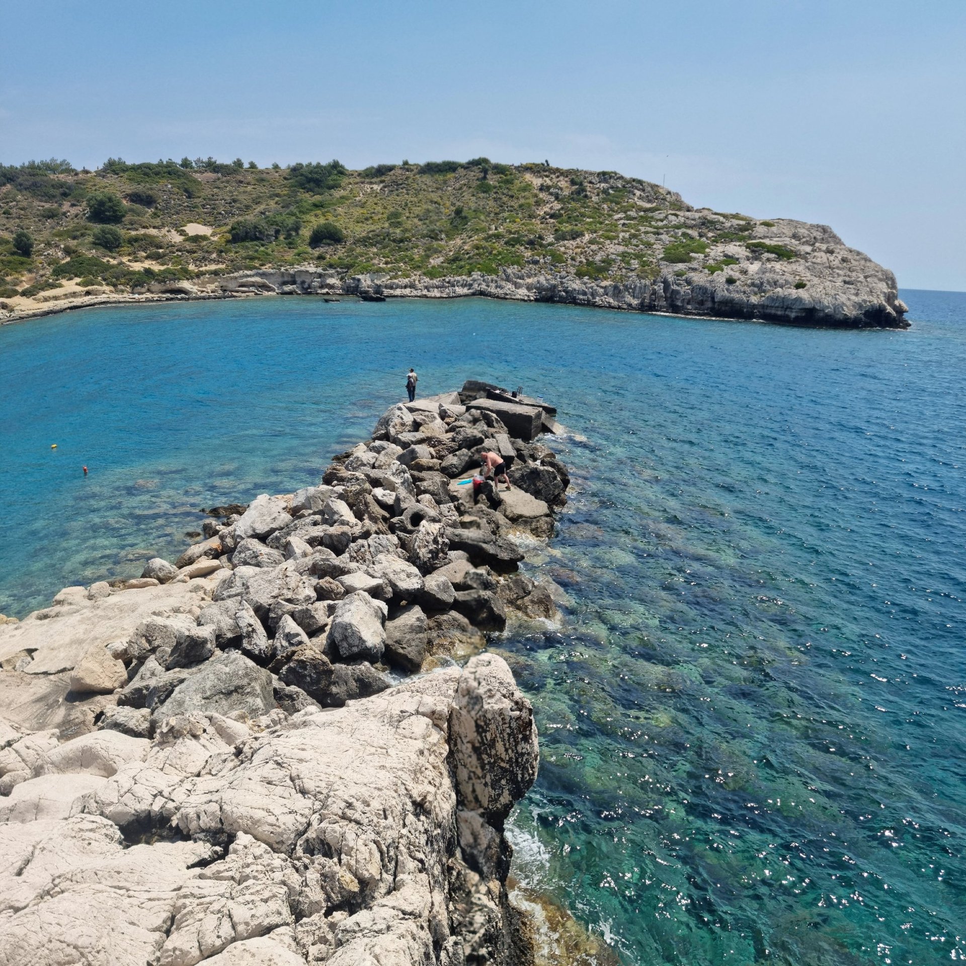 People relax and explore on a rocky breakwater in a beautiful, clear blue Mediterranean cove, perfect for snorkeling and