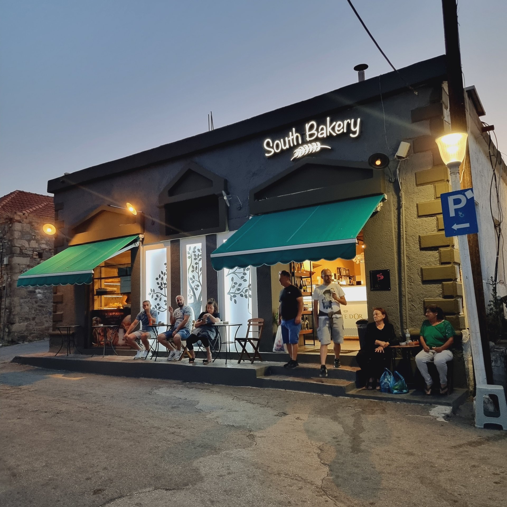 Customers relax outside South Bakery, a modern storefront with green awnings and outdoor seating at twilight.