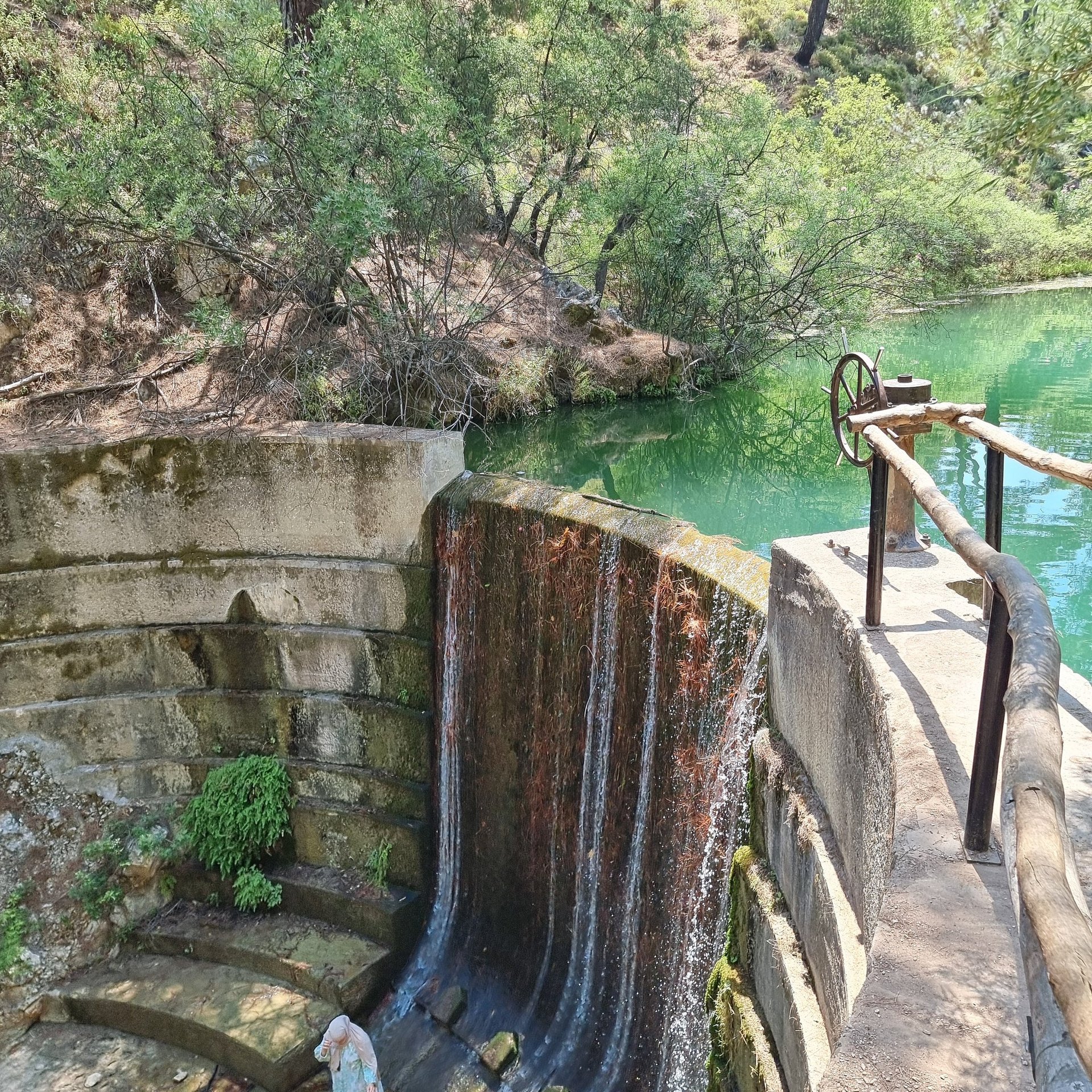 A small concrete dam and waterfall flowing into a turquoise pond surrounded by lush green trees.