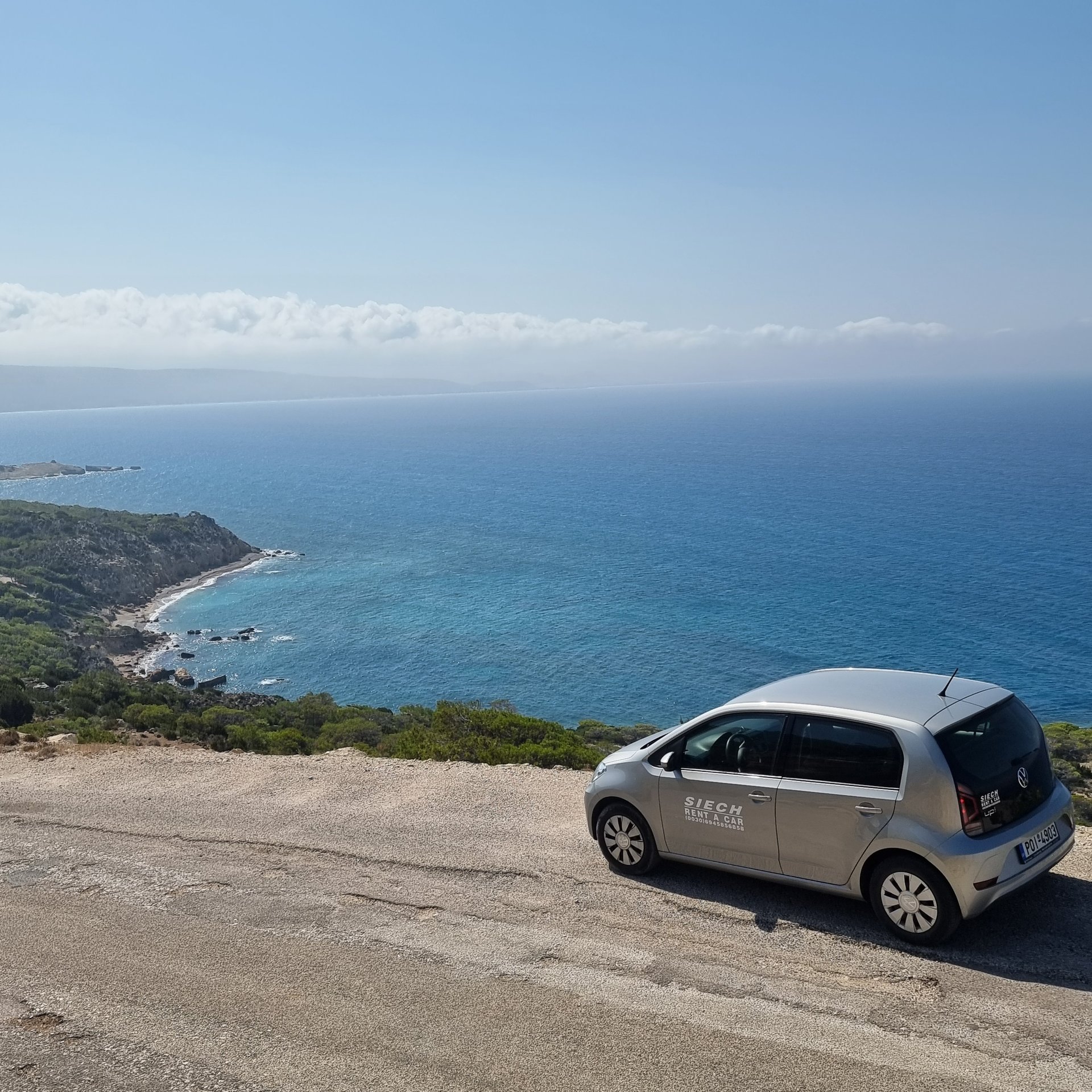 A silver compact rental car parked on a scenic coastal overlook with a view of the blue Mediterranean sea.