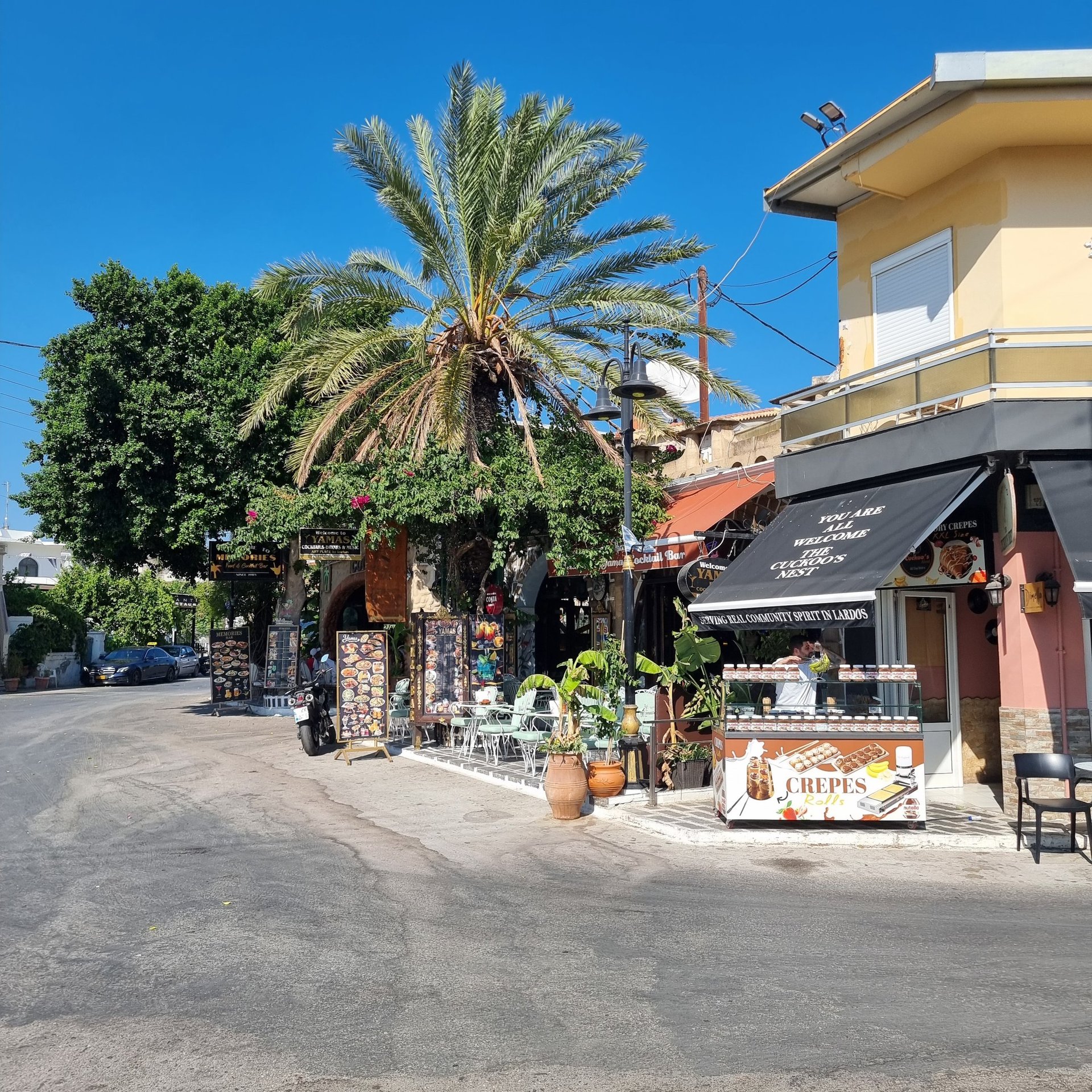Sunny street view of a Lardos village creperie and cafe with outdoor seating and palm trees in Rhodes, Greece.