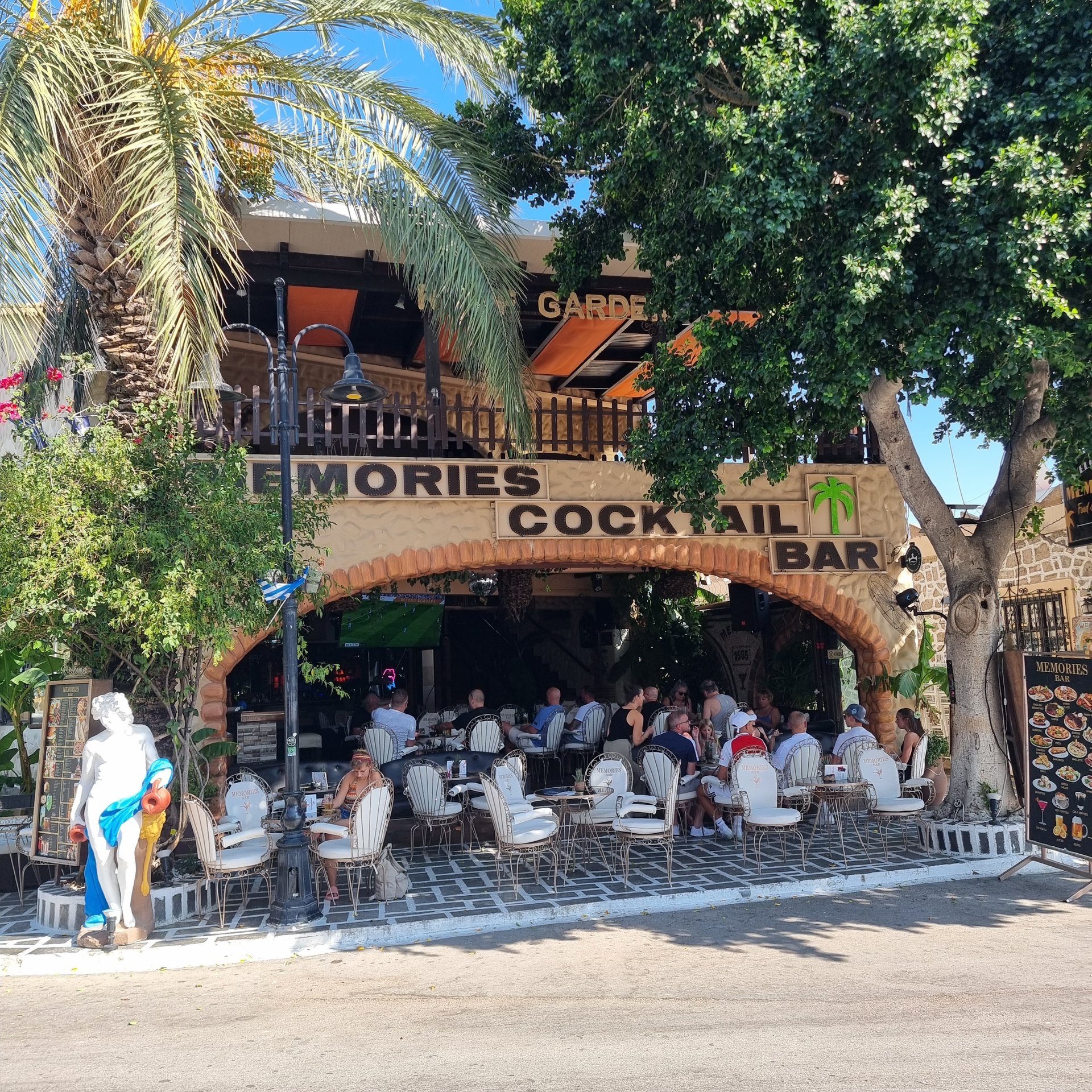 Patrons enjoy drinks at the outdoor Memories Cocktail Bar terrace under palm trees in Greece.