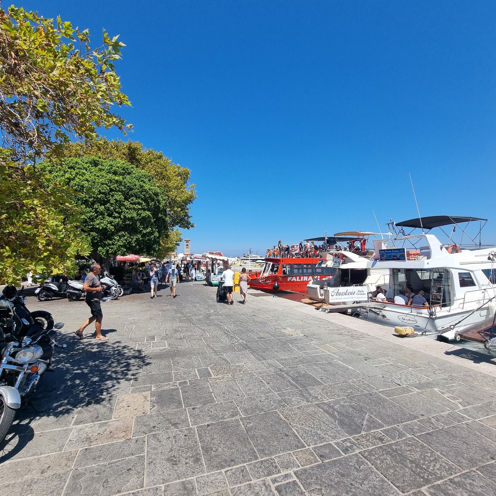 Crowded Mandraki Harbor in Rhodes Greece with tourist boats moored along the stone promenade under a blue sky.