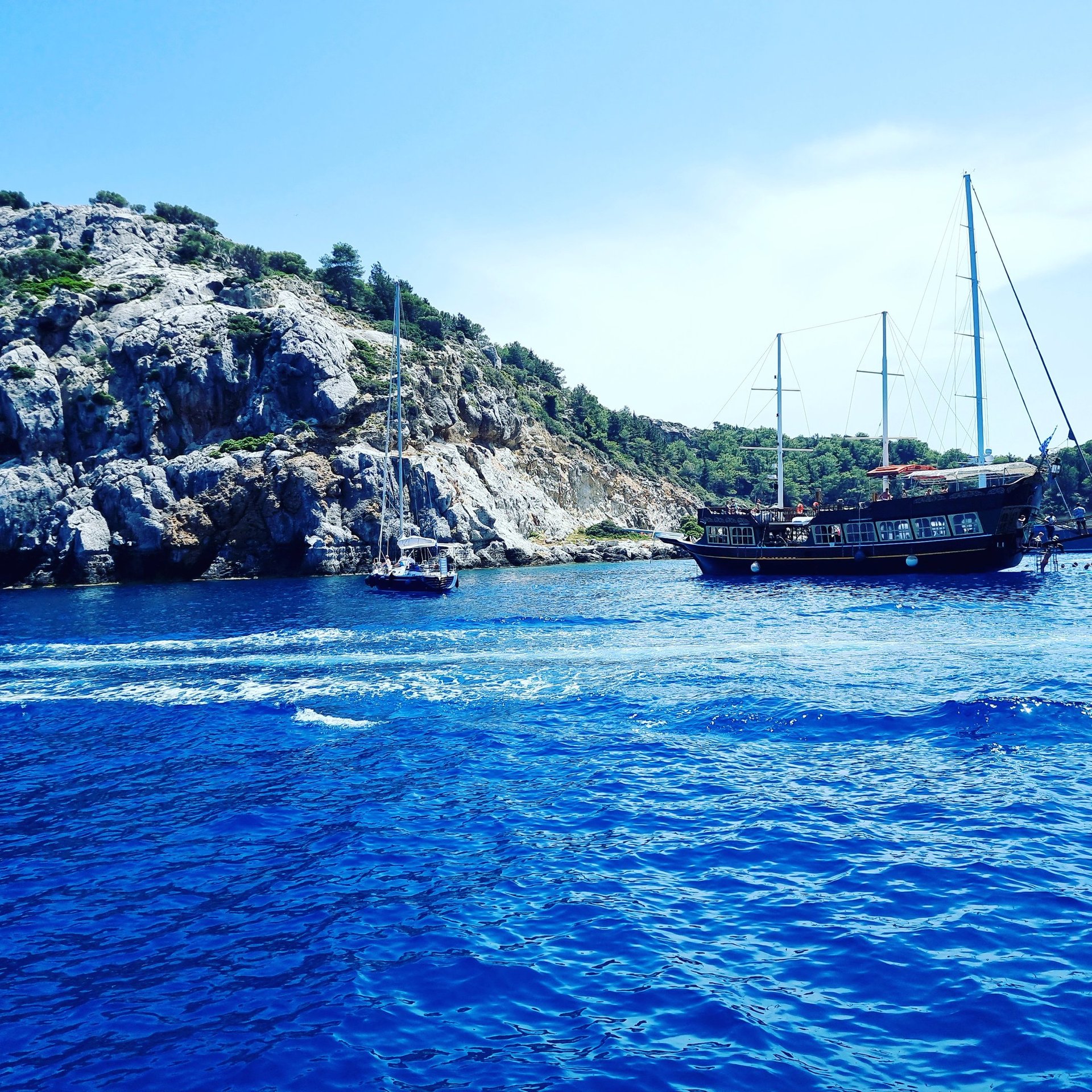 A wooden pirate ship and a sailboat anchor in the bright blue water of a Mediterranean bay.