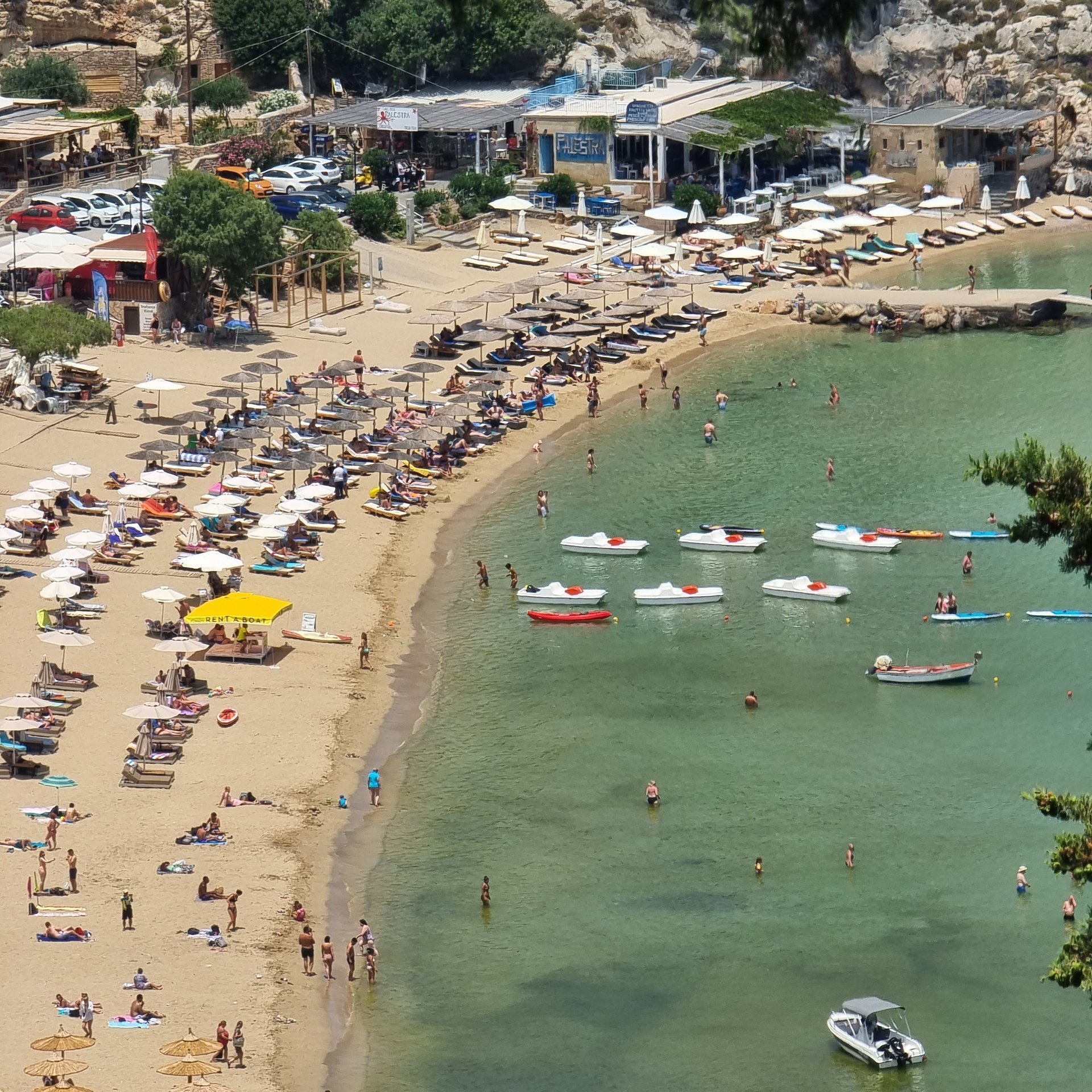 Aerial view of a crowded Mediterranean beach with rows of umbrellas and boats in the turquoise sea.