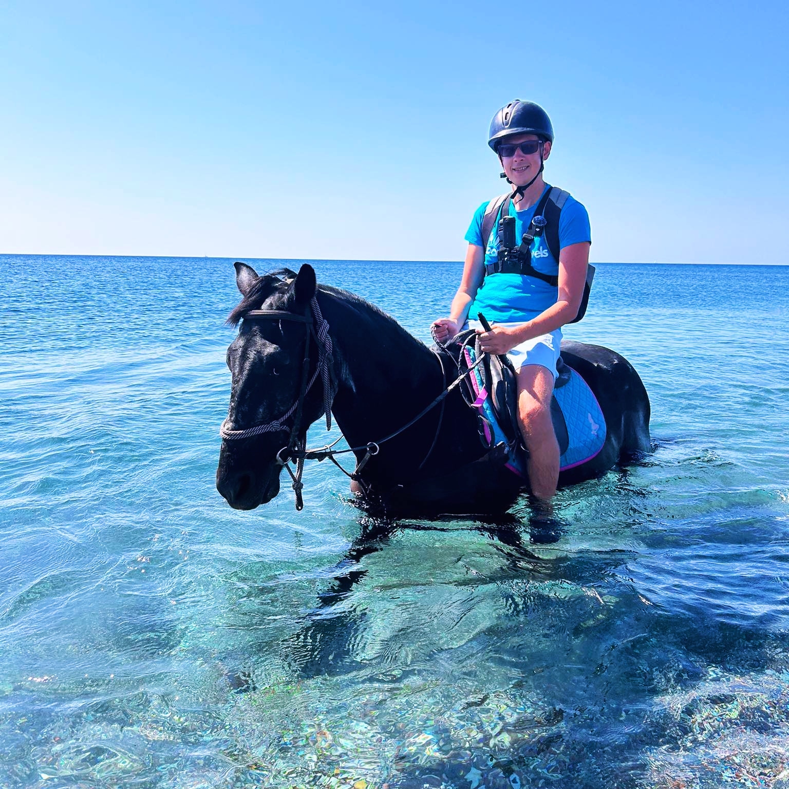 looneytravels riding a black horse through clear blue ocean water during a summer beach horse trek.