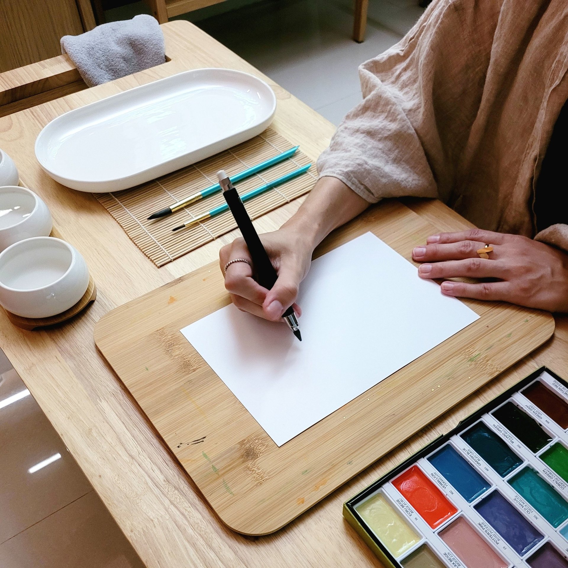An artist drawing on white paper with a black pen, surrounded by watercolor paints and brushes.