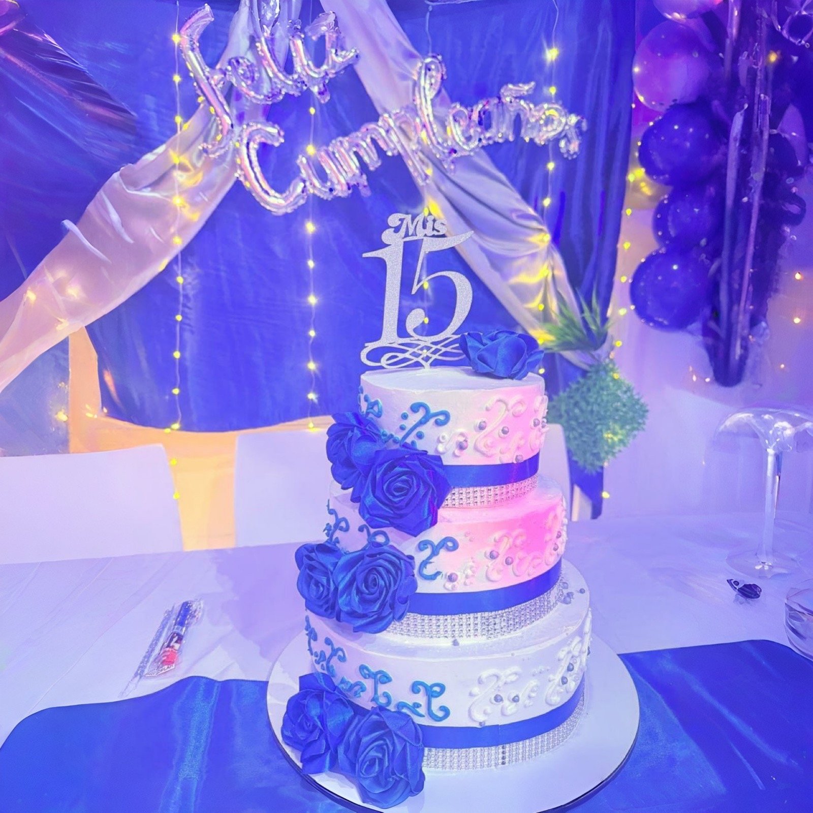 Elegante pastel de quinceañera de tres niveles con rosas azules y detalles plateados, con un adorno 