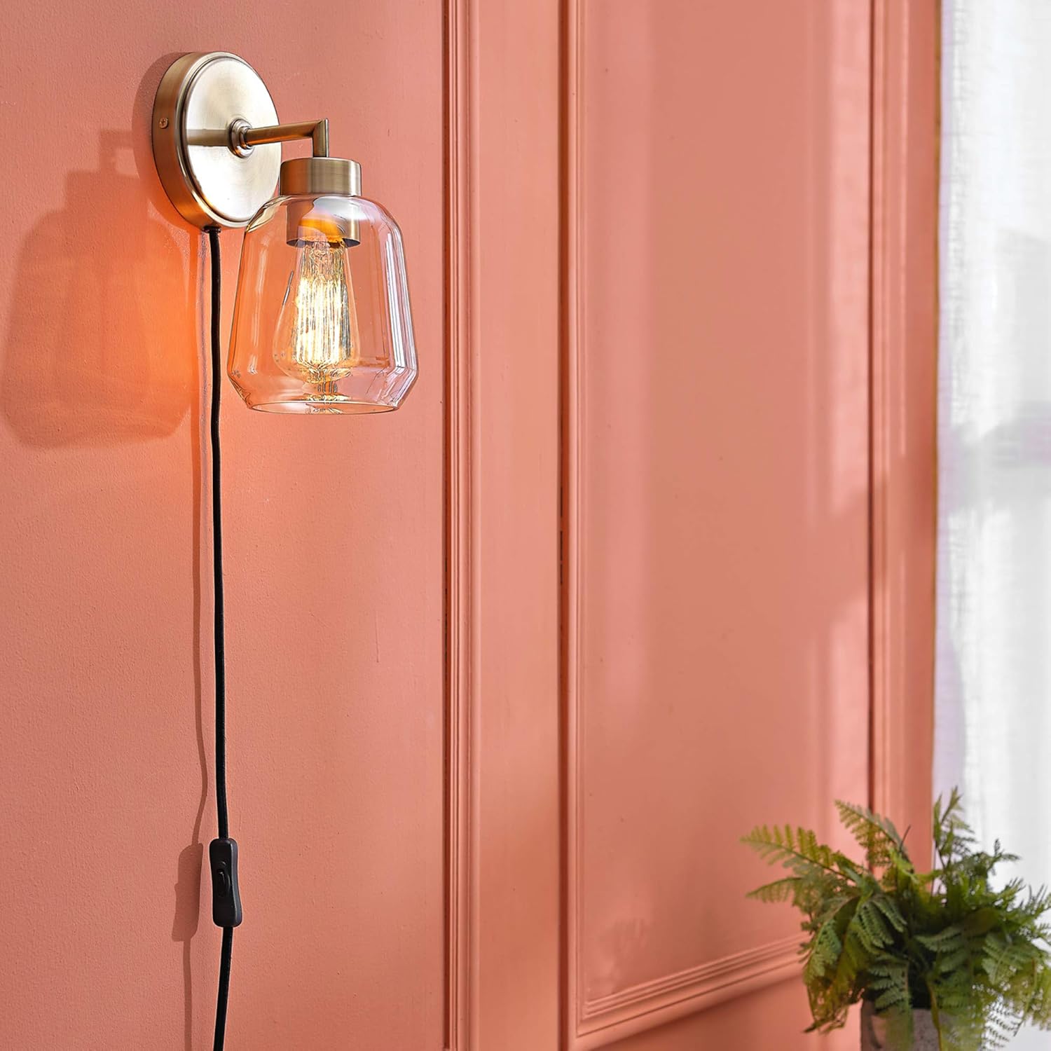 Modern brass wall sconce with a clear glass shade and Edison bulb mounted on a salmon pink wall.