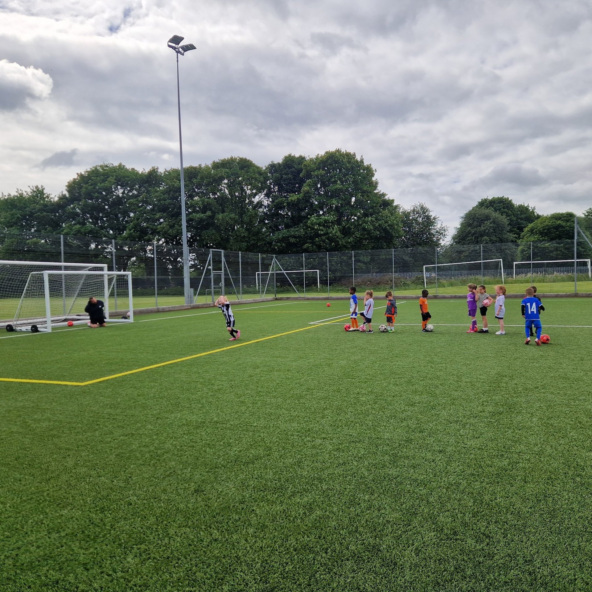 Young children practice footbal drills on a green artificial turf field with goals and footballs.