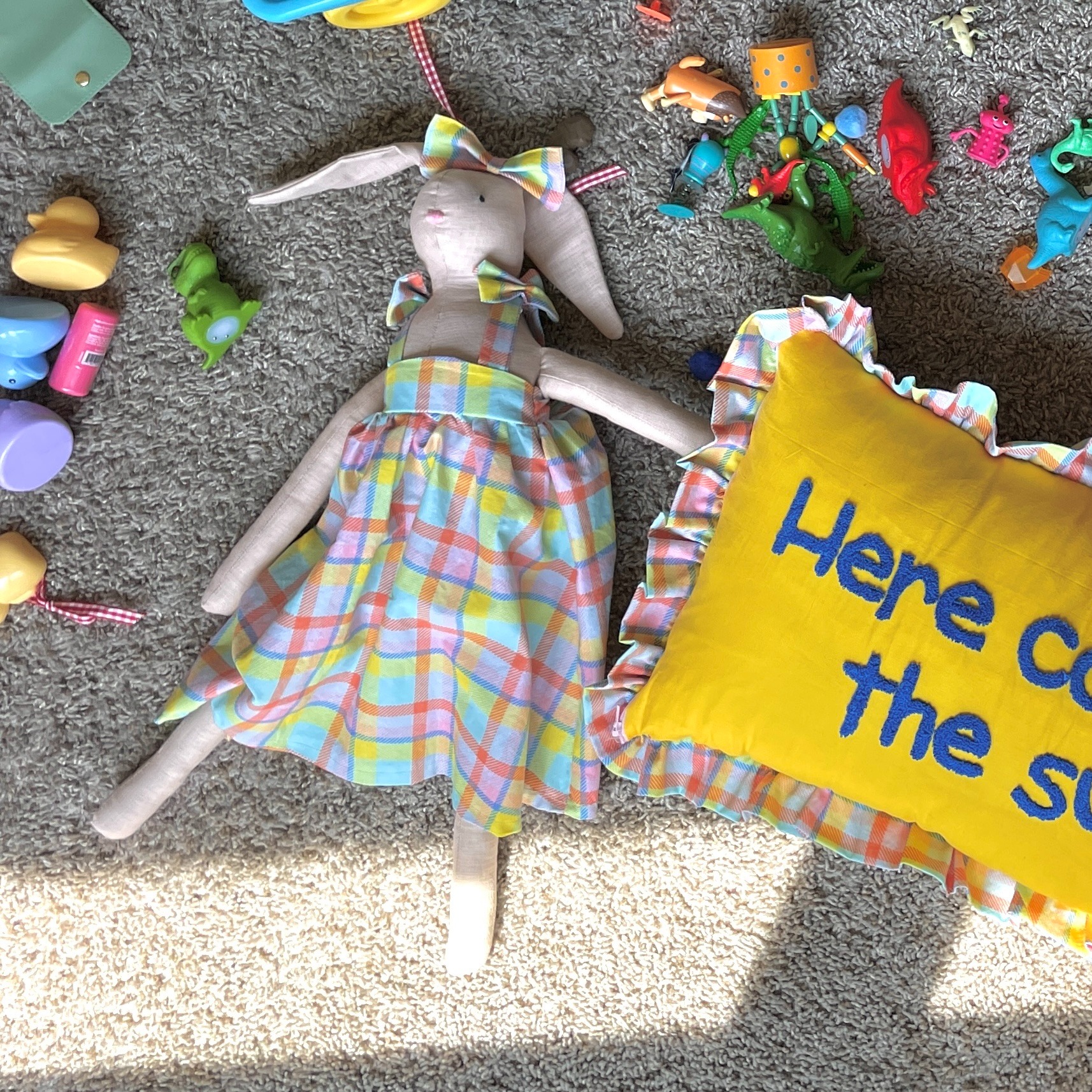Handmade cloth bunny doll in a plaid dress lying on a carpet with kids toys and a yellow pillow.