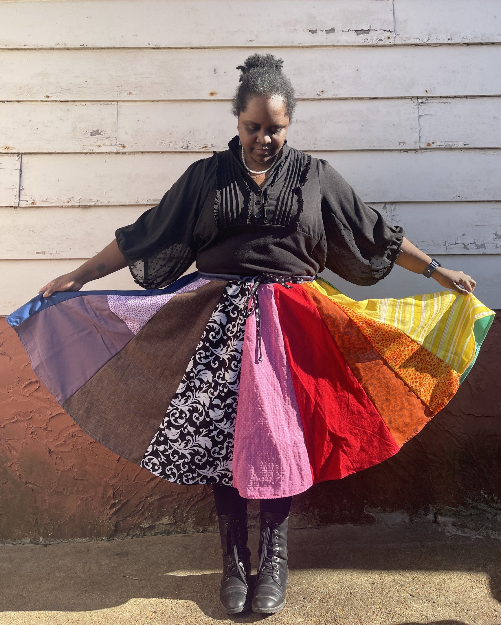 a black woman wearing a black shirt and rainbow circle skirt standing against a wall