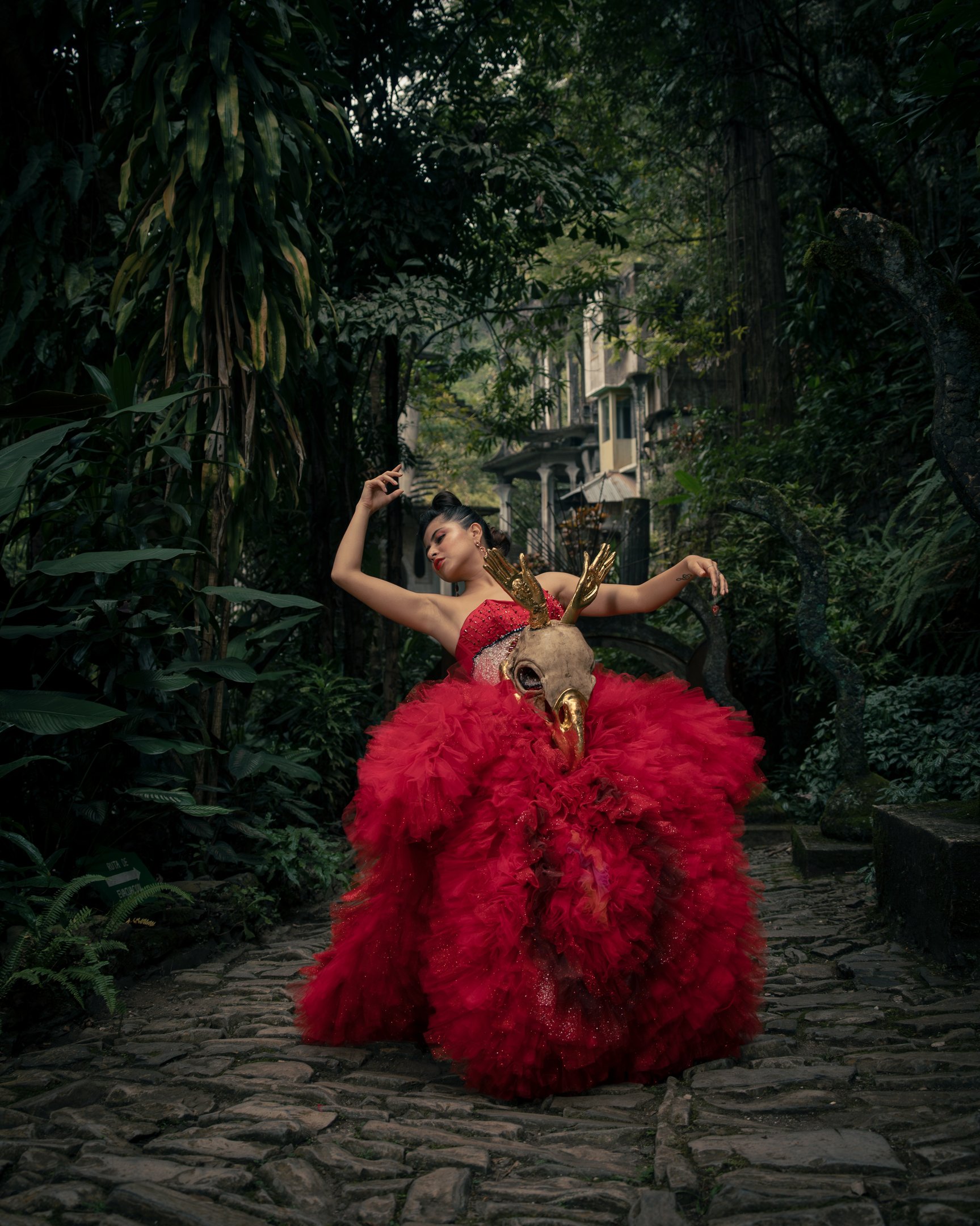 A woman in a dramatic red tulle gown and gold deer skull mask poses in a lush tropical garden.