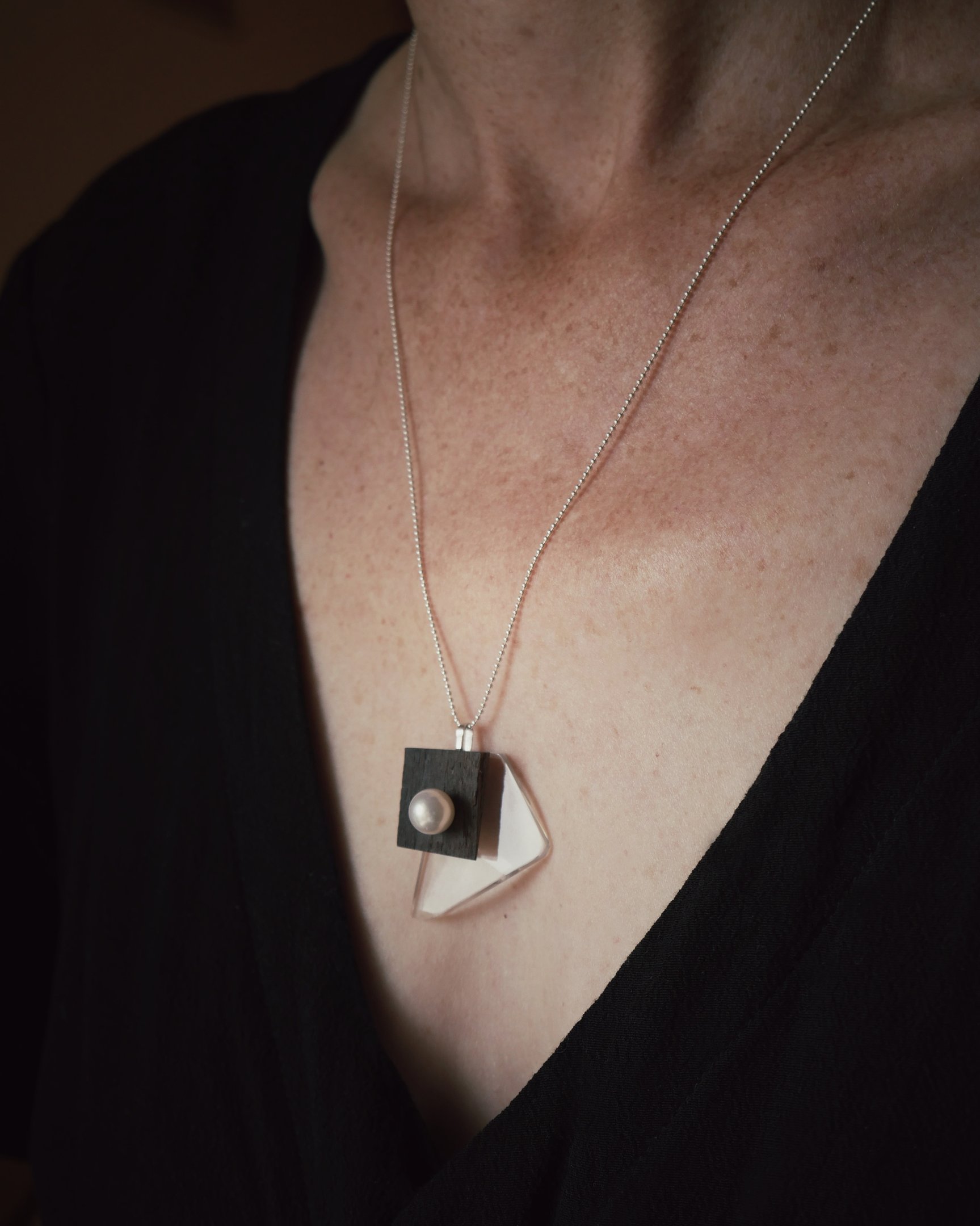 A modern geometric pendant necklace with silver chain, ebony wood, acrylic, and a pearl.