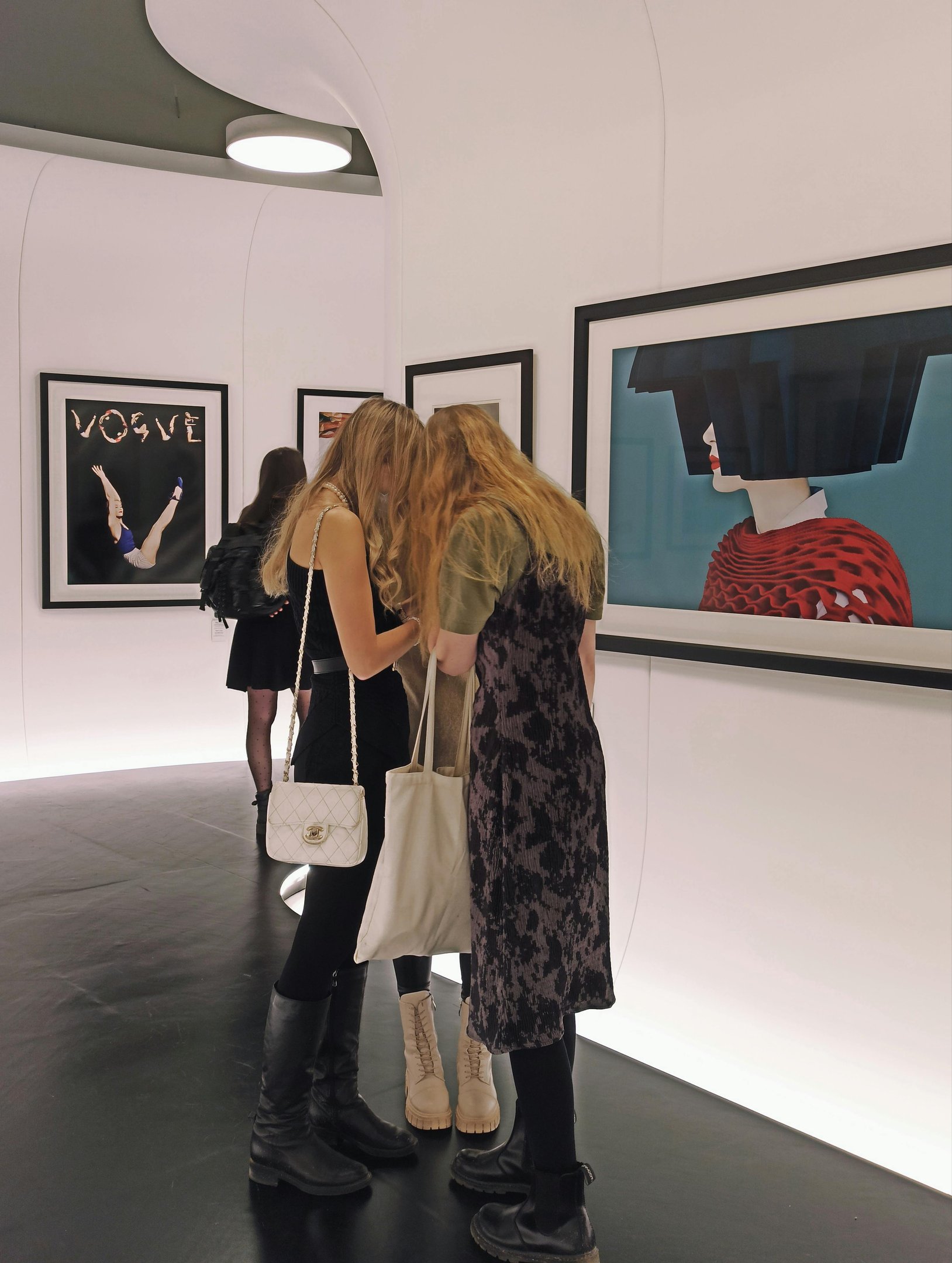 Visitors view artworks at a public exhibition; two women embrace in front of framed photographs.