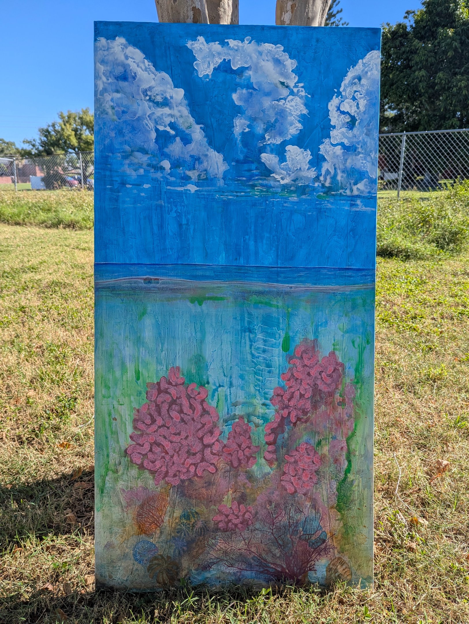 A color dense painting of a coral reef with a clouded blue sky above rendered in gloss acrylics on linen by Kathy LaFollett.
