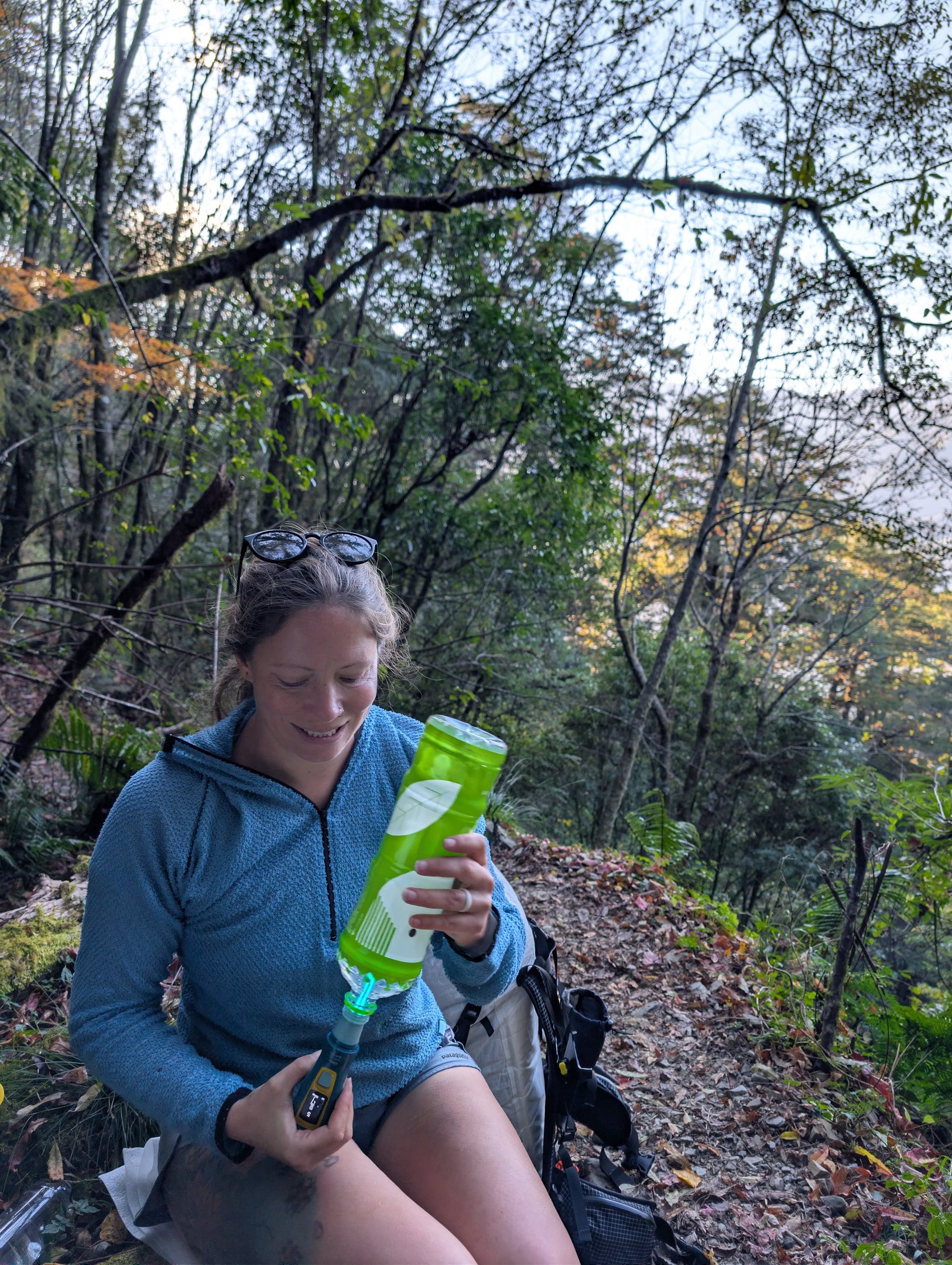 A female hiker filters water using a Katadyn Ultra UV filter into a green bottle on a forest trail.