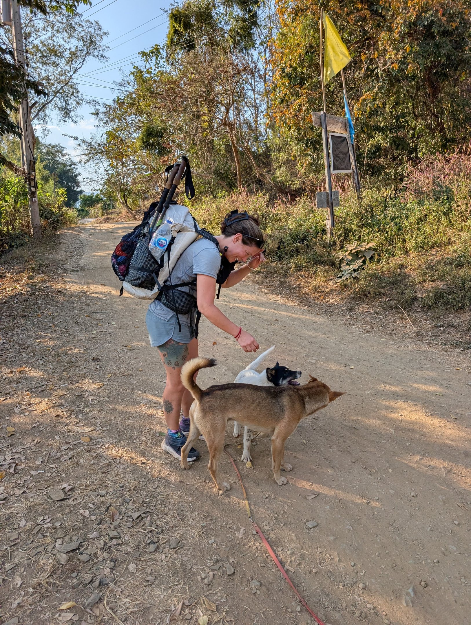 A female hiker with a Hyperlite backpack petting two dogs on a sunny dirt road in Norther Thailand..