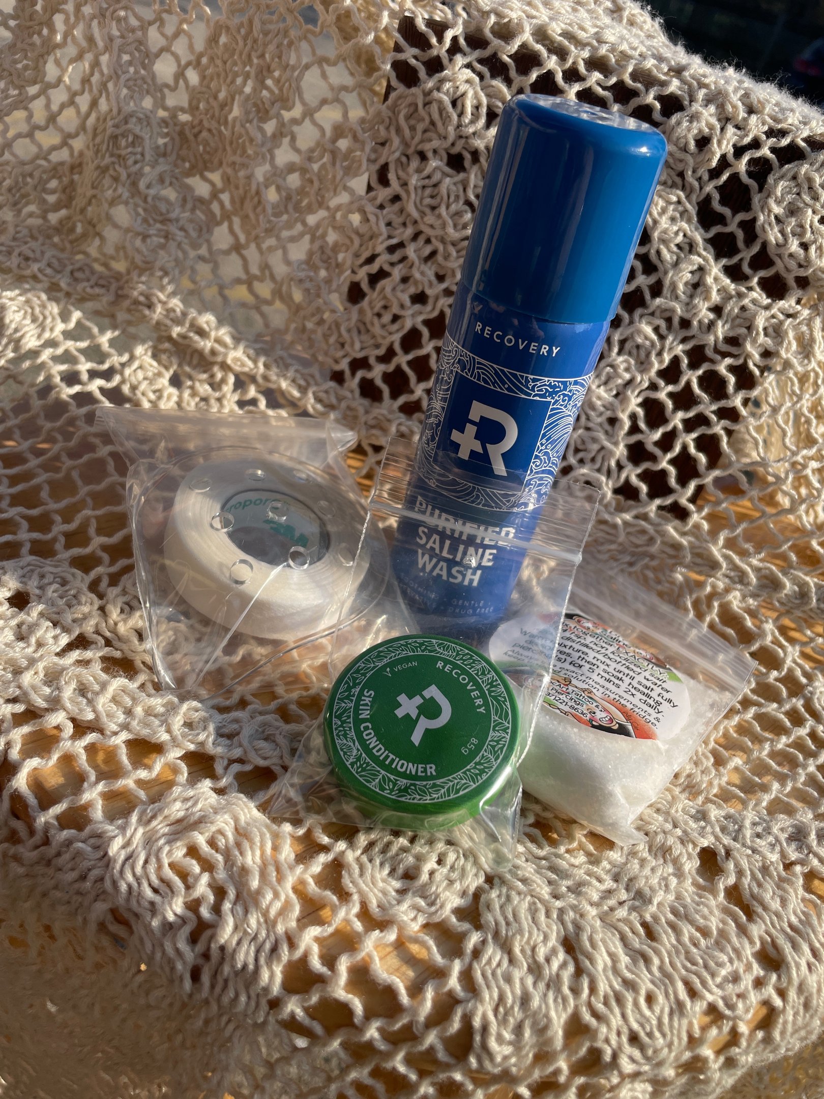 Piercing aftercare kit flatlay: Recovery brand saline spray & skin salve, protection patch + medical tape, & bag of sea salt.