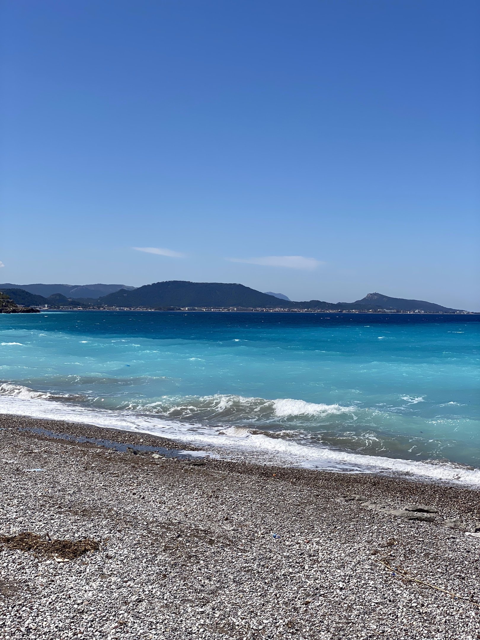 Pebble beach with turquoise Mediterranean waves and mountains under a clear blue sky.