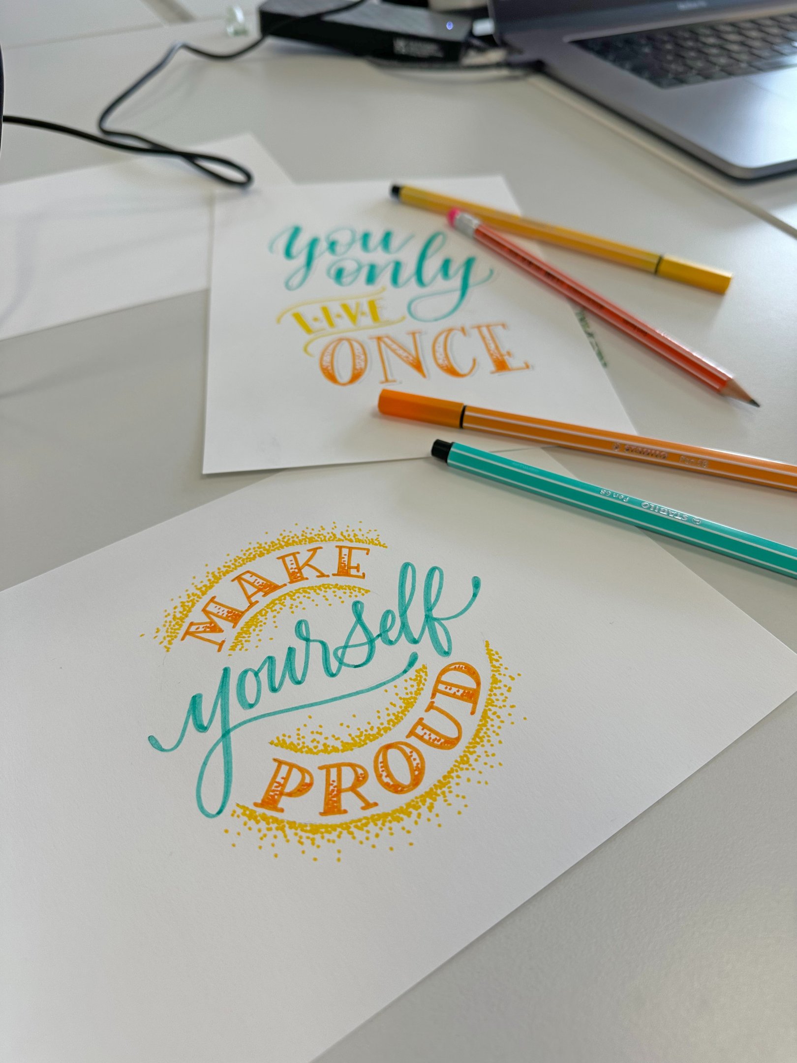 Hand-drawn lettering art on cards with motivational quotes and colorful pens on a desk.