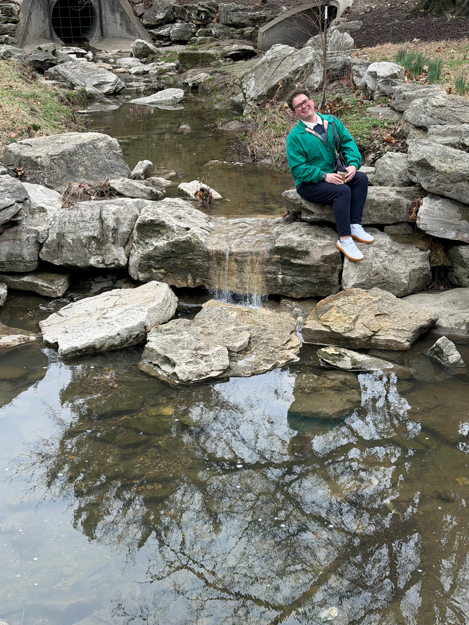 A person in a green jacket sits by a rocky stream and small waterfall in a scenic park.