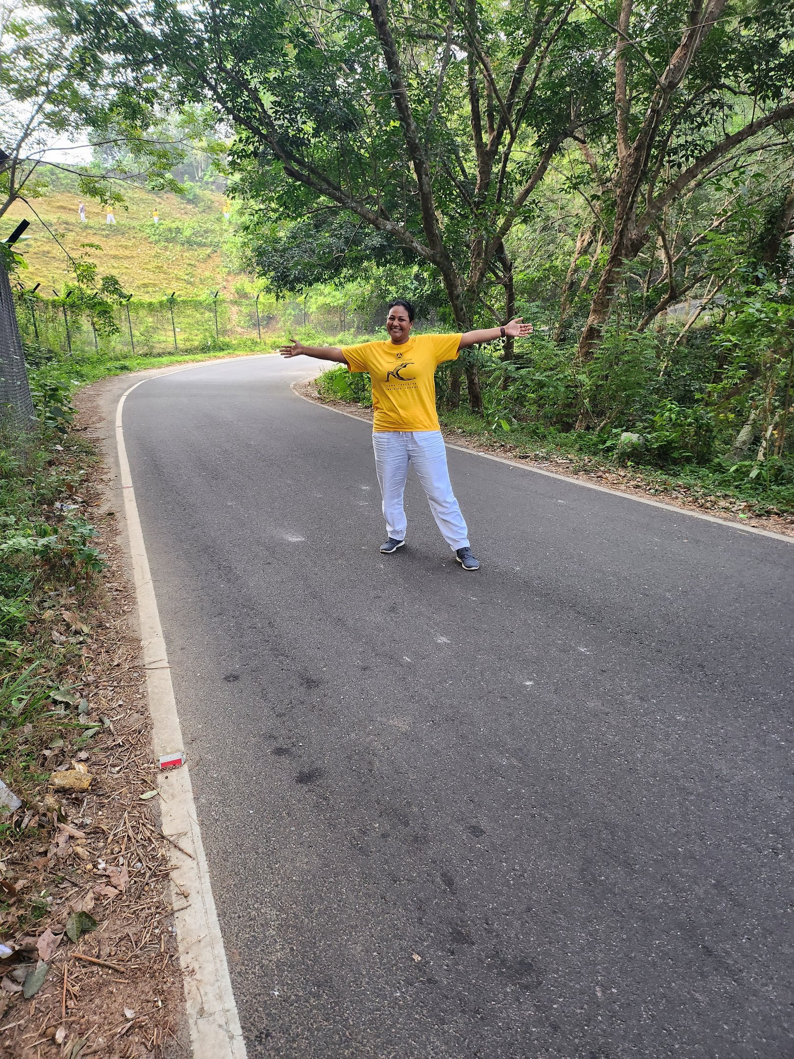 A smiling woman in a yellow shirt stands with arms open on a winding paved road through a lush green forest.