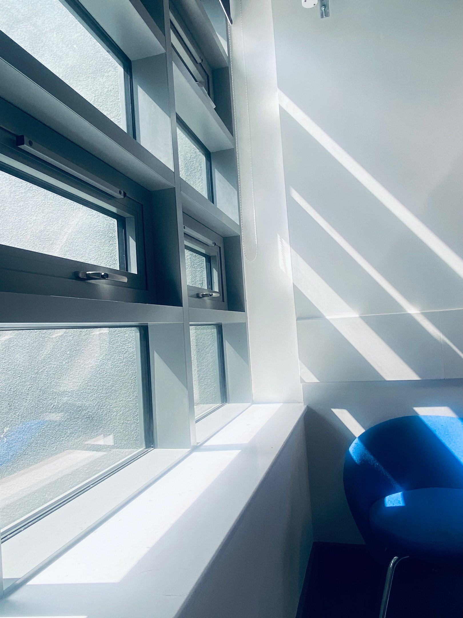 Modern office window with natural sunlight shadows casting over a blue chair and white wall.