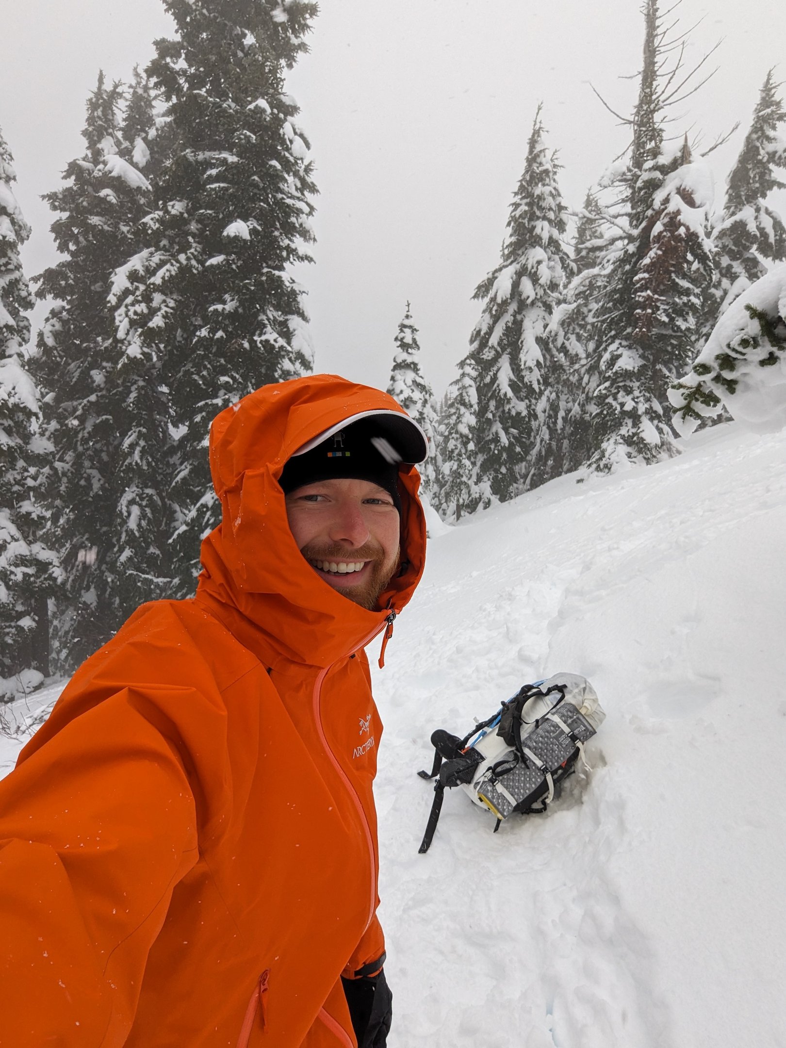 A smiling man wearing an orange Arc'teryx jacket with a Hyperlite backpack lying in the snow.