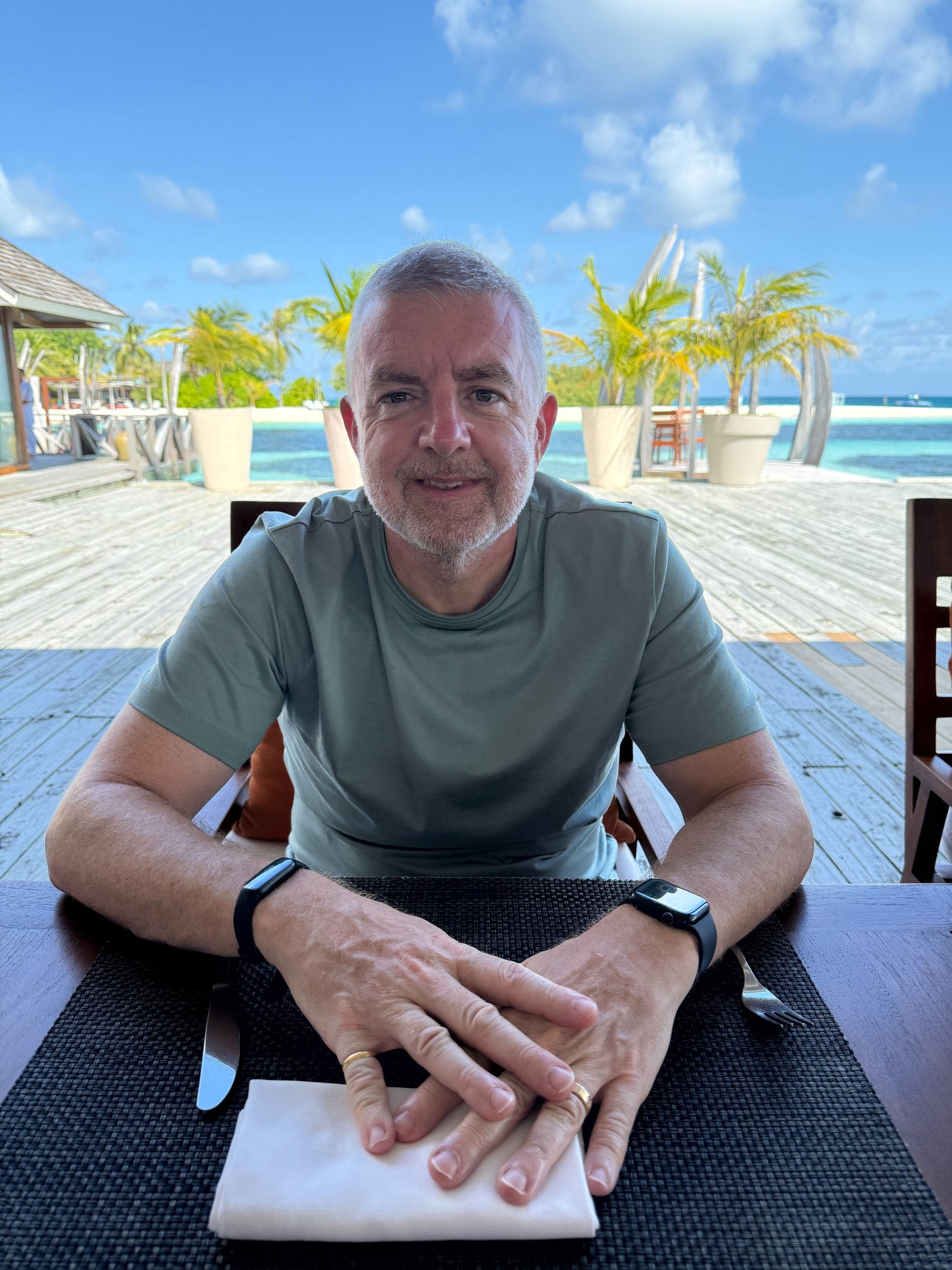 "Person seated at outdoor table in tropical resort with palm trees and lounge chairs nearby