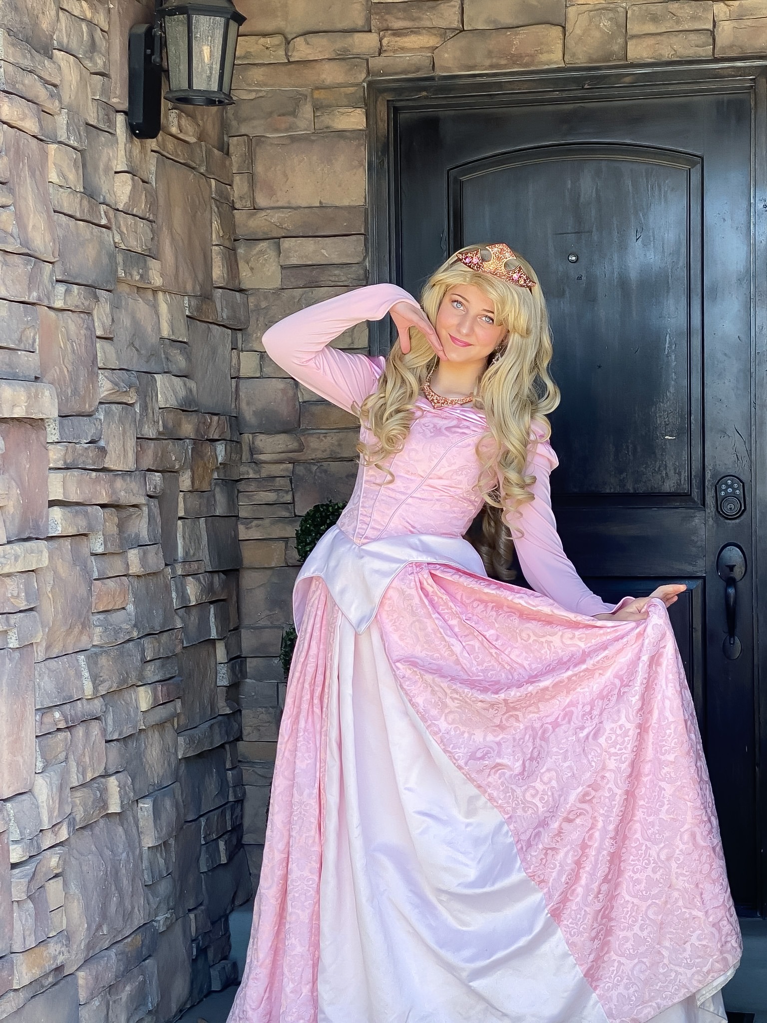 A blonde princess performer in a pink Aurora dress and crown poses by a stone wall for a birthday party.