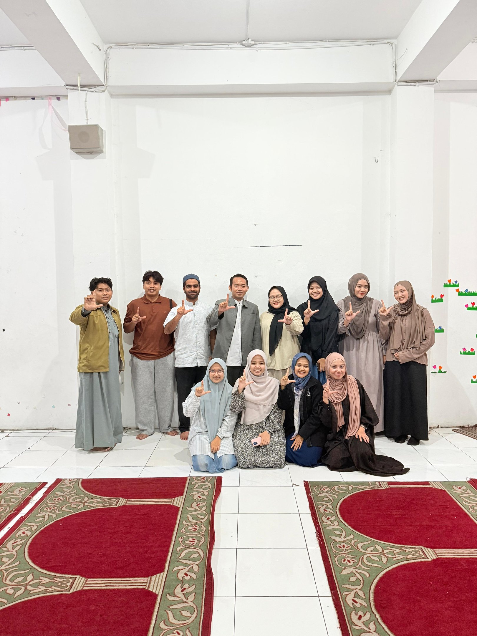 A group of Muslim students in a prayer room making hand gestures on red patterned rugs.