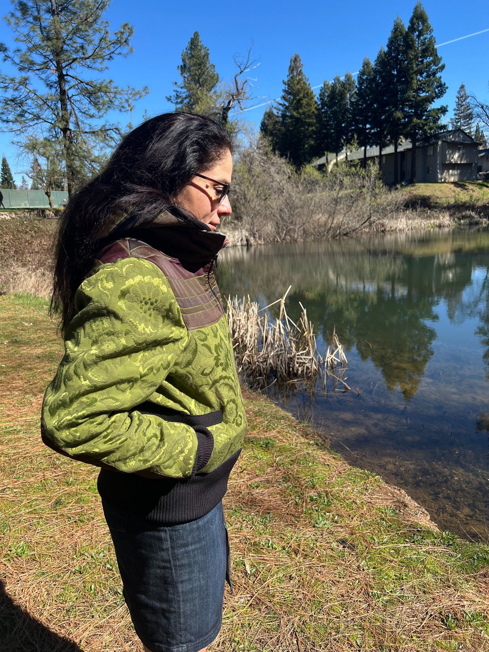 a woman in a green jacket and jeans standing in front of a pond