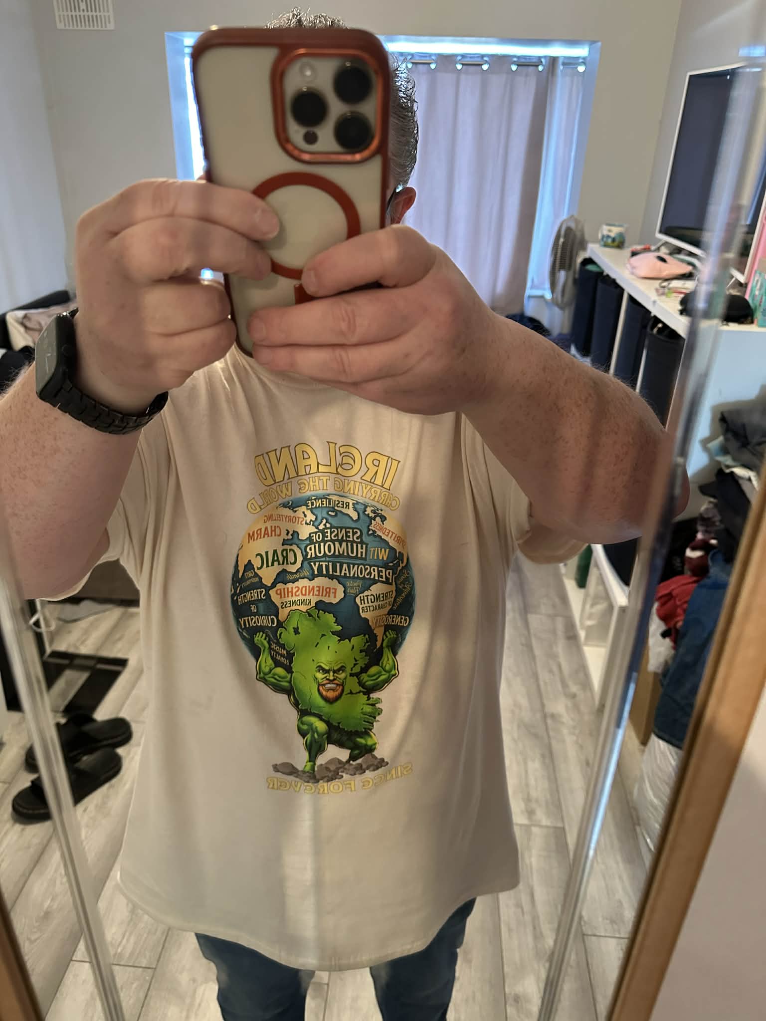A man takes a mirror selfie wearing an off-white graphic t-shirt featuring a green Irish mythological figure.