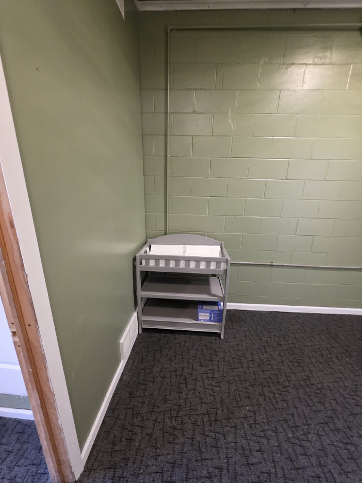 A gray wooden baby changing table with shelves stands against a green cinder block wall in a nursery room.